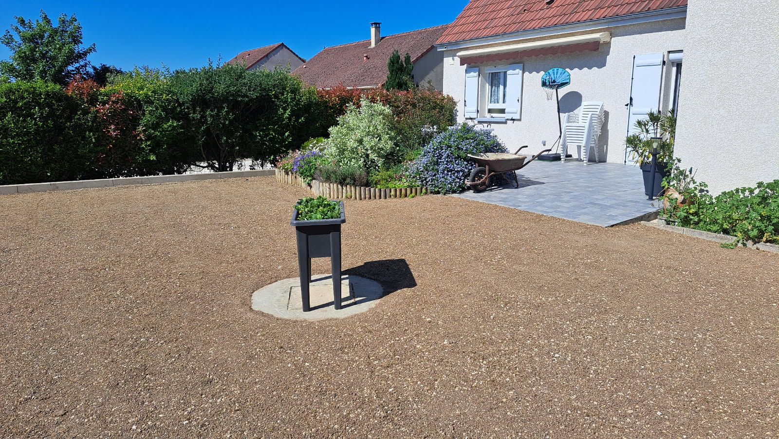 Cour de gravier avec parterre de fleurs, jardinière et maison claire au toit rouge sous un ciel lumineux.