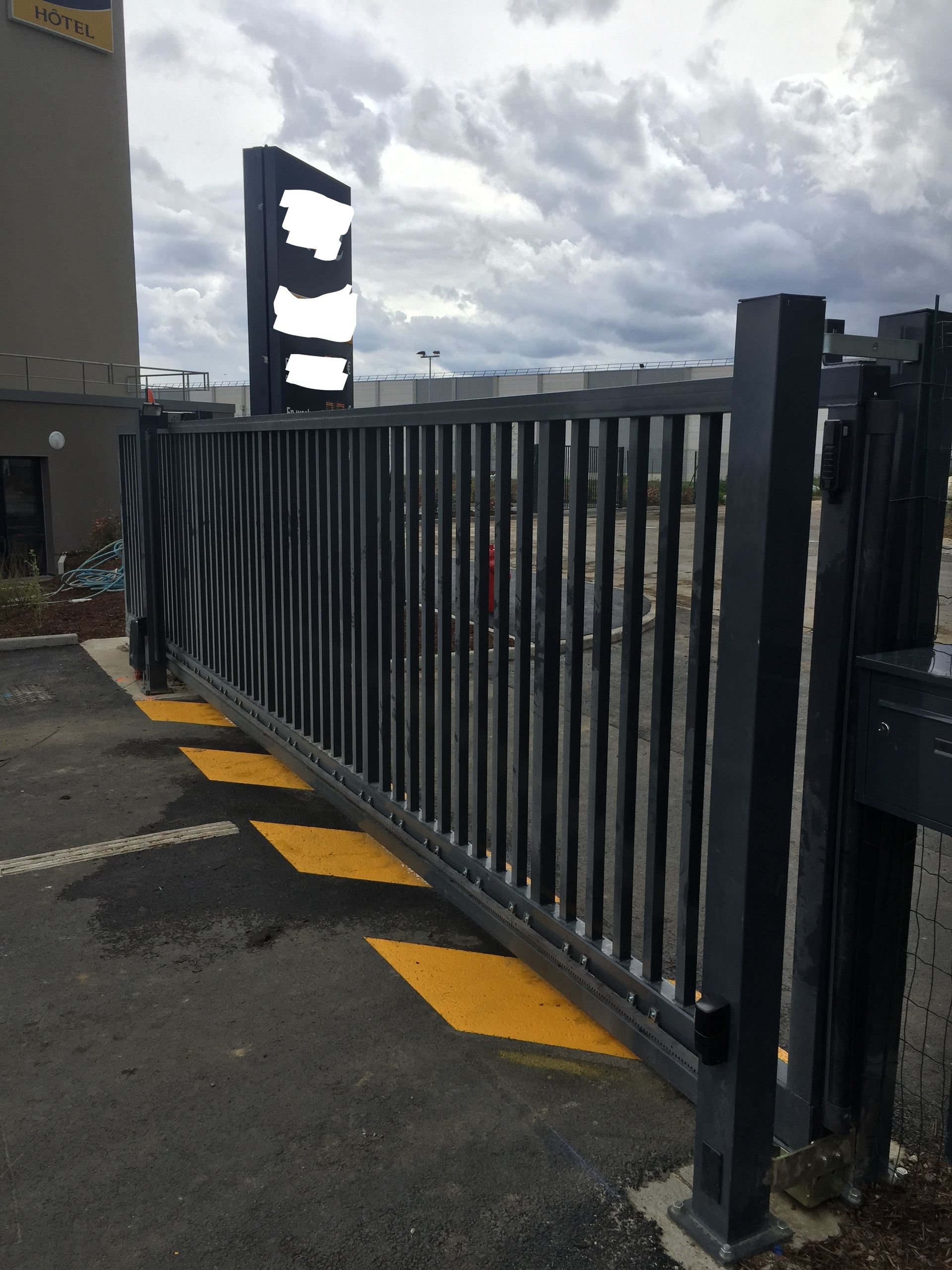 Un portail métallique noir bloque l'entrée d'un parking, avec un panneau en arrière-plan.