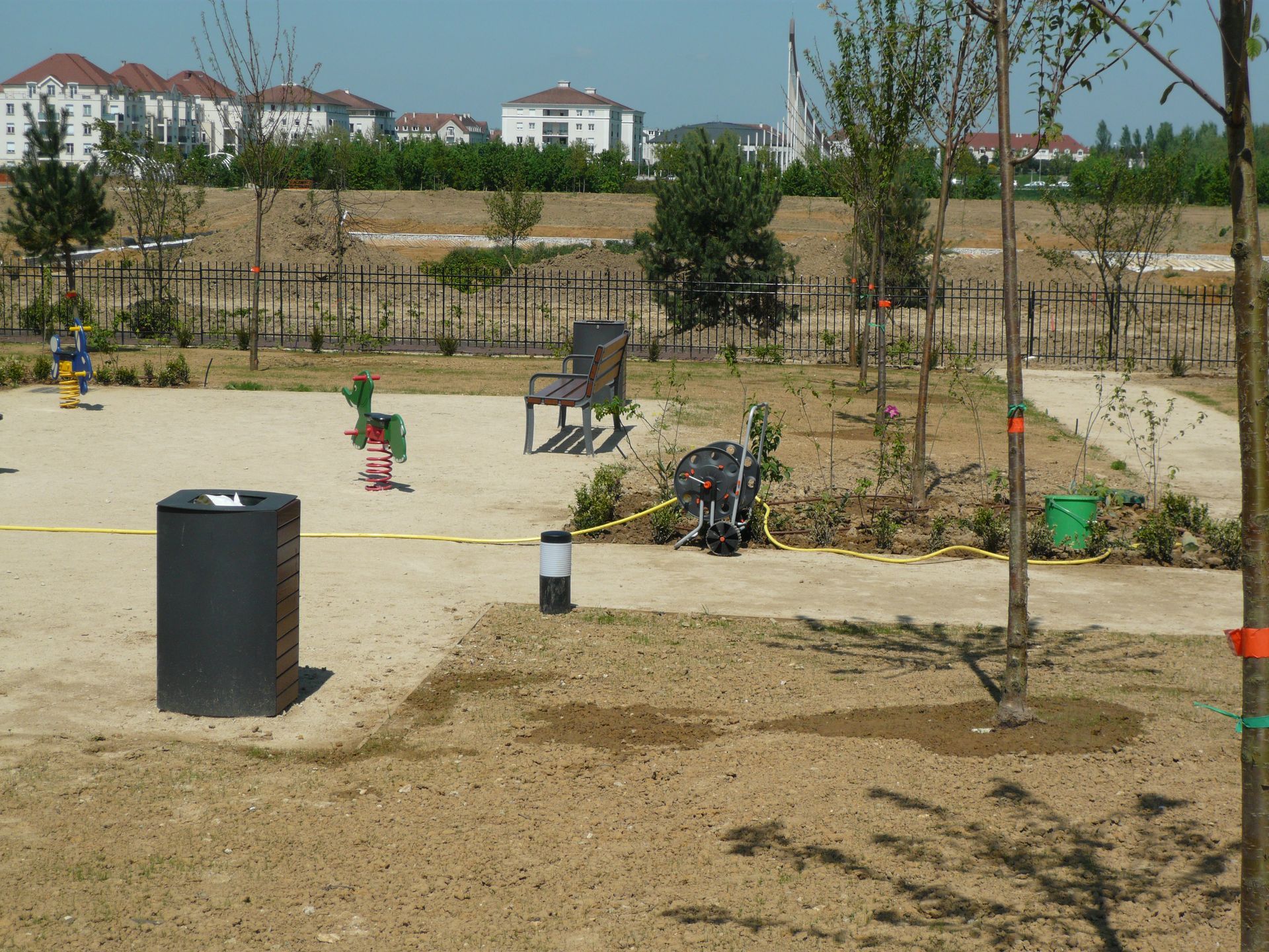 Aire de jeux avec du sable, des arbres, une chaise et une poubelle, avec des immeubles d'appartements en arrière-plan.