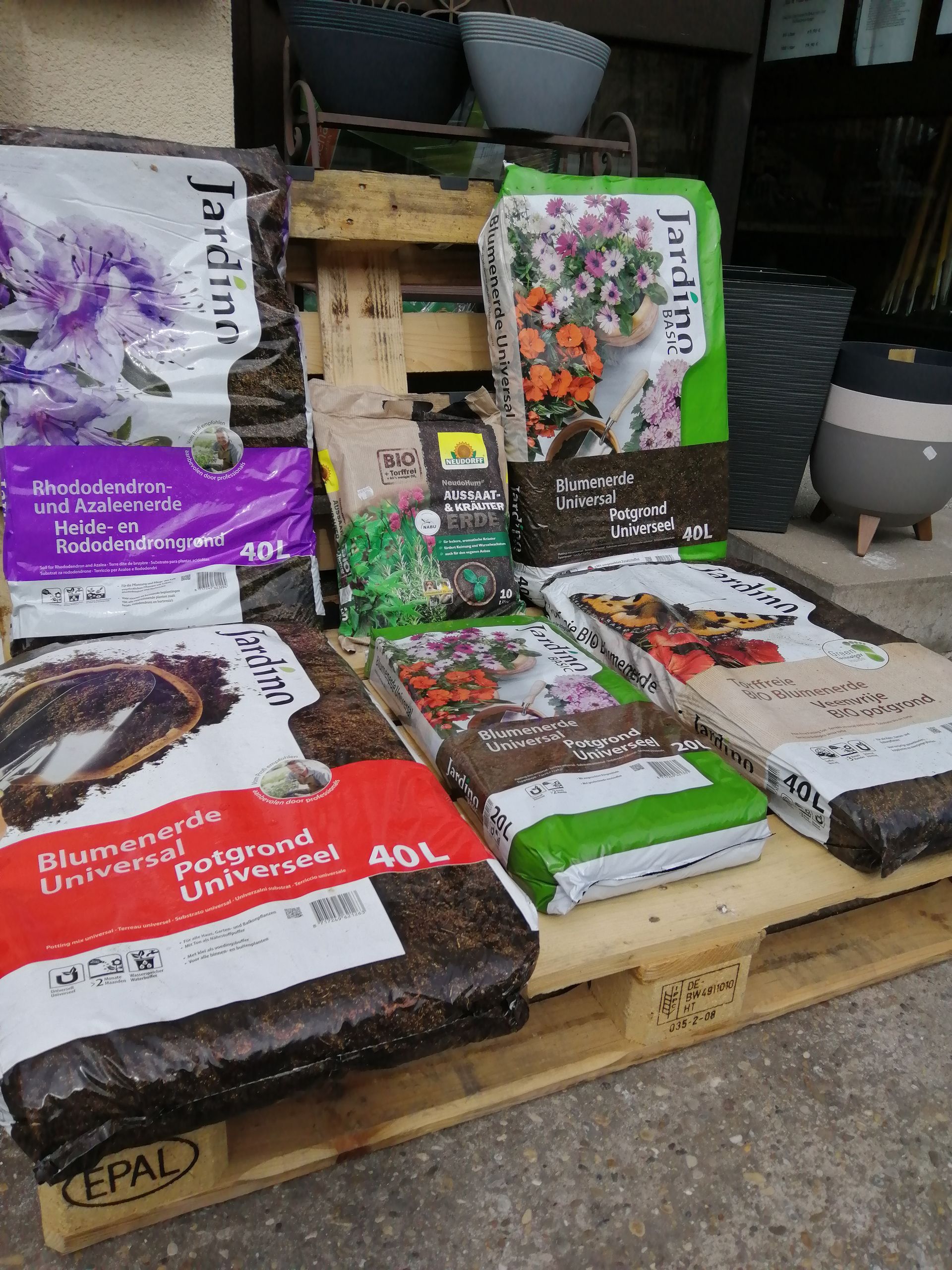 Säcke mit Blumenerde verschiedener Marken und Farben auf einer Holzpalette in einem Gartencenter.