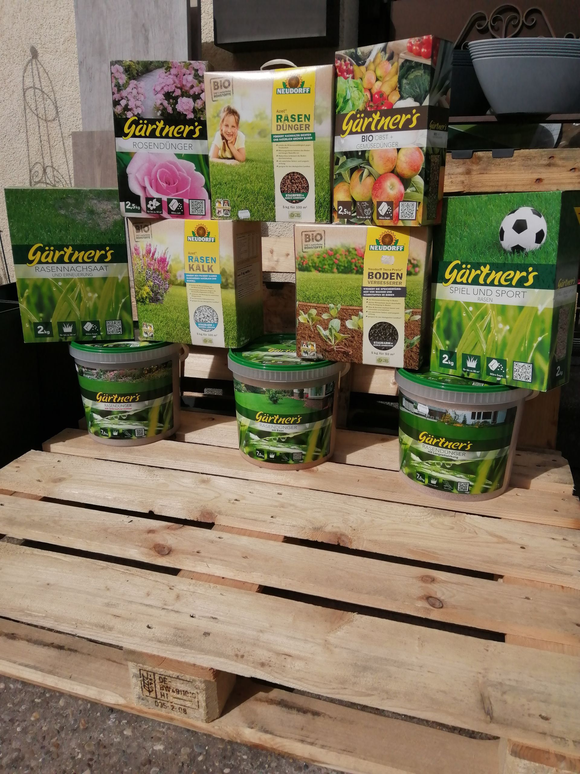 Präsentation von Pflanzendüngern auf einer Holzpalette. Kisten und Eimer mit verschiedenen Düngemitteln in grüner, weißer und gelber Verpackung.