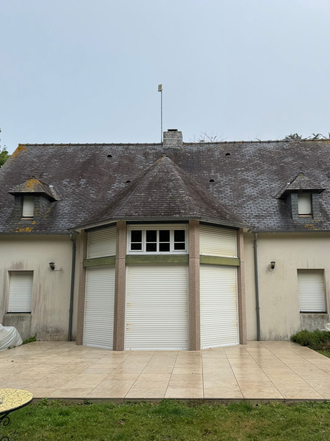 Maison en pierre avec garage central et deux volets latéraux, donnant sur une terrasse carrelée.