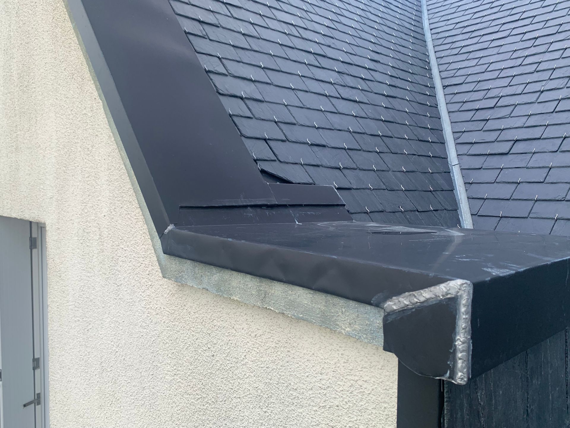 Bord de toit avec bardeaux foncés, solin noir et descente pluviale blanche sur un angle de bâtiment.