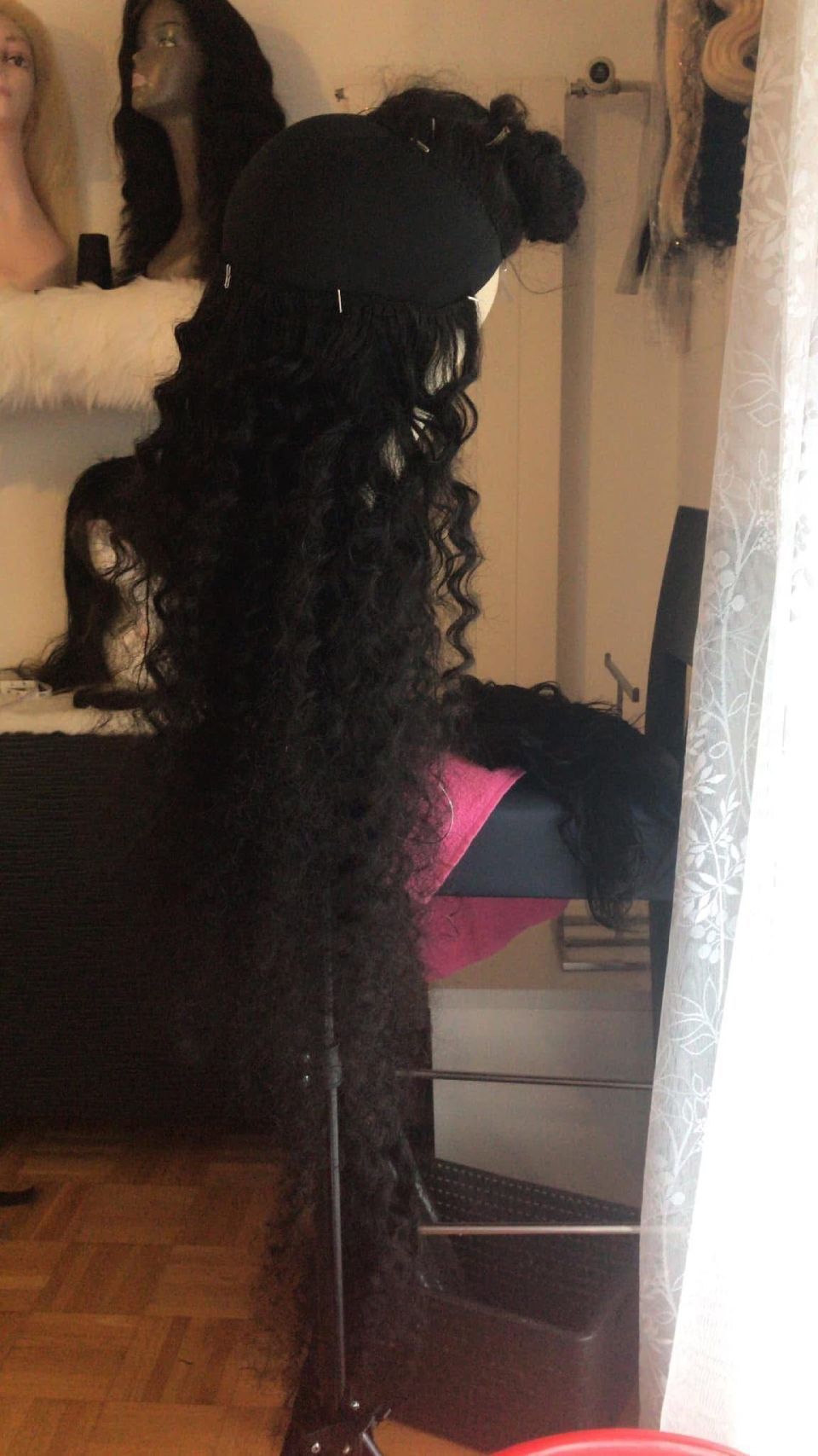 Schwarze Lockenperücke auf einem Perückenständer in einem Raum mit anderen Perücken und einem durchsichtigen weißen Vorhang.