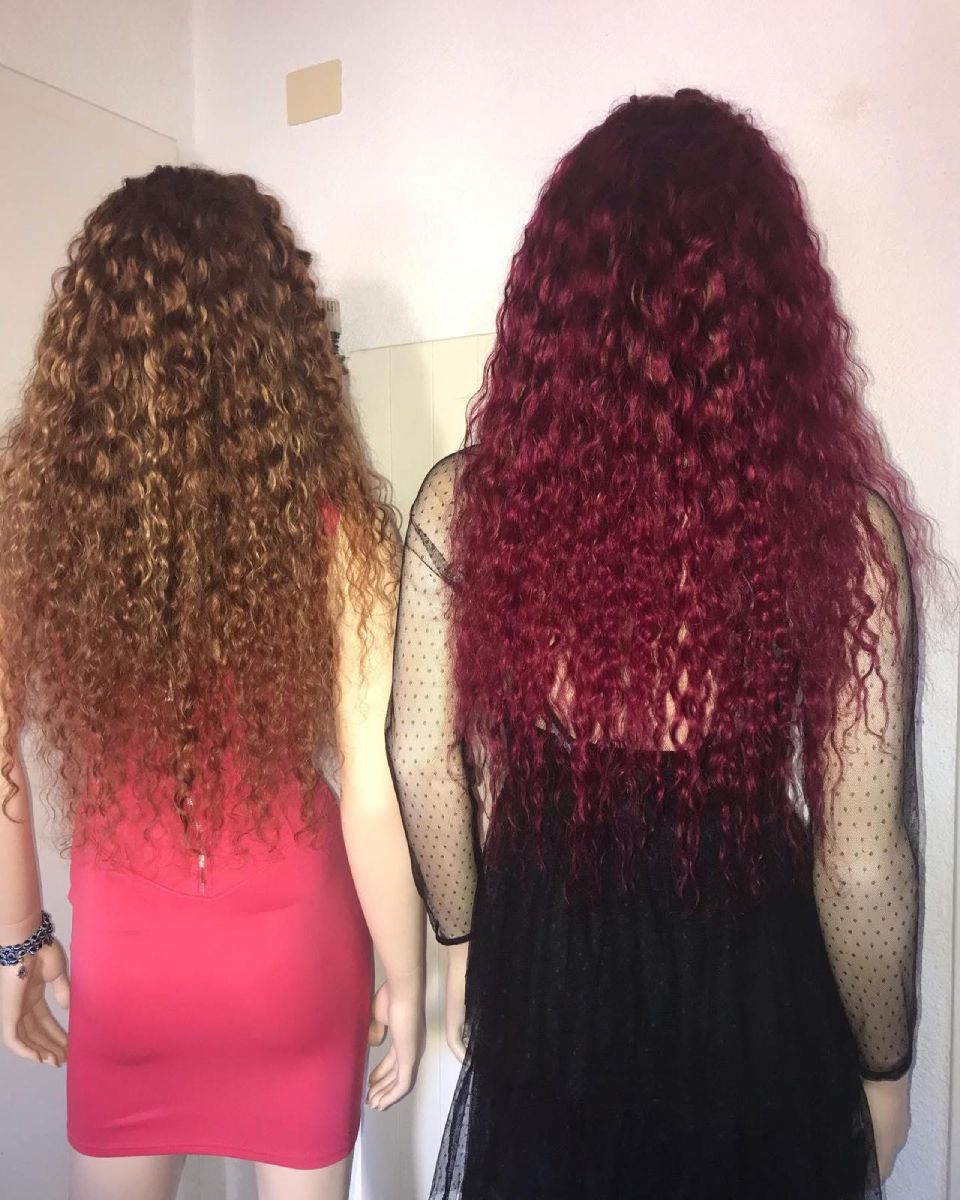 Zwei Schaufensterpuppen mit langen, lockigen Perücken, eine rot, eine braun, tragen Kleider und stehen im Innenbereich.