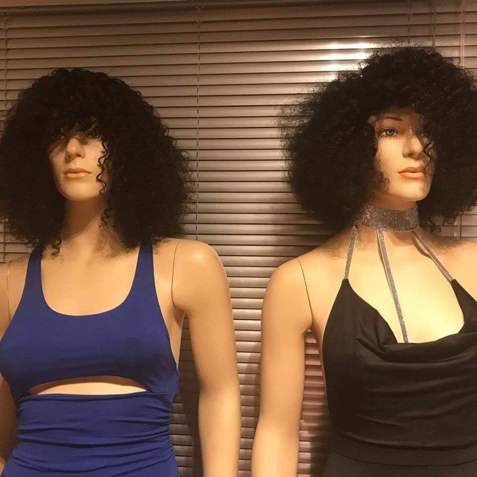 Zwei Schaufensterpuppen mit schwarzen Afros, eine im blauen Kleid, eine im schwarzen Kleid, lehnen an Jalousien.