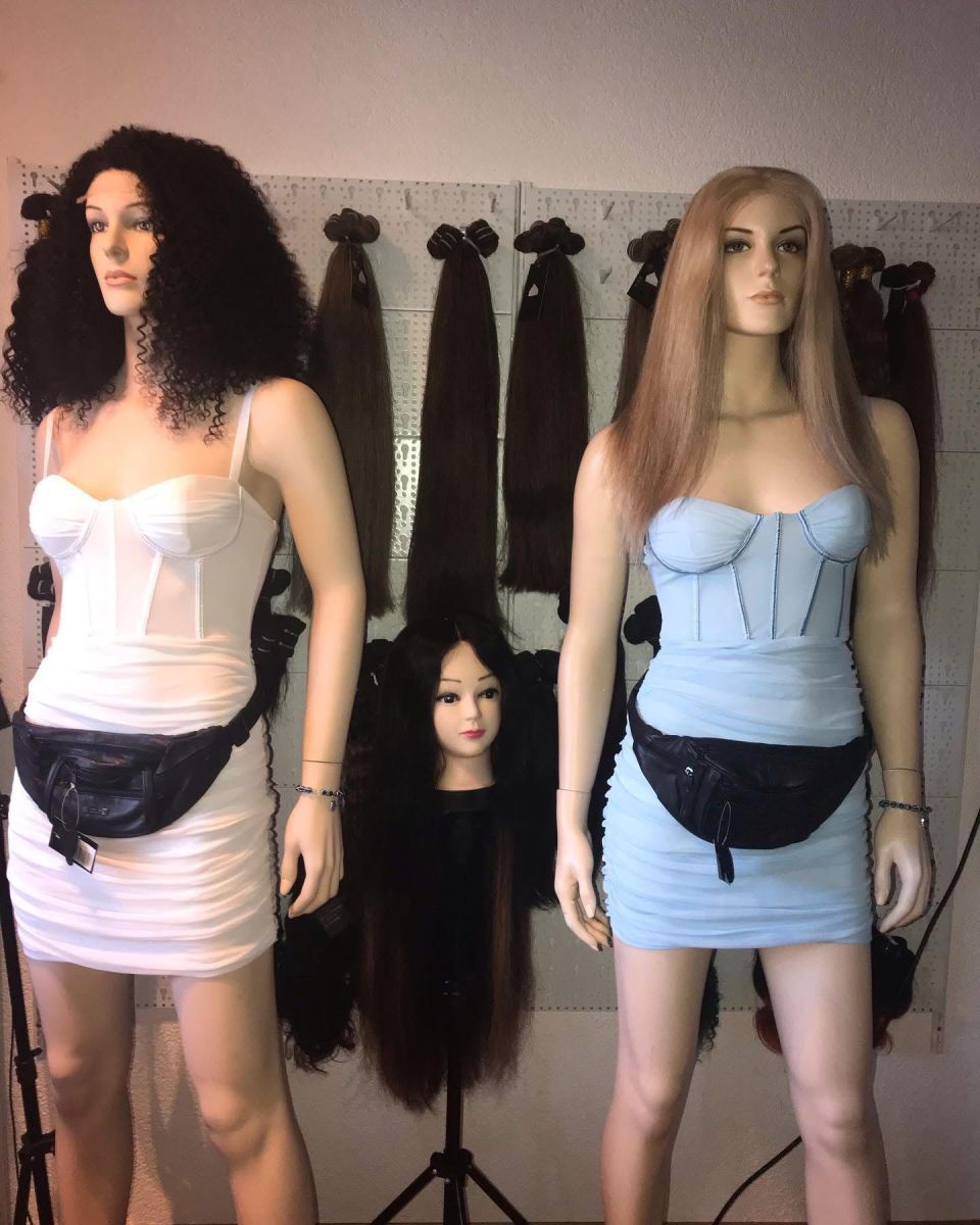 Zwei Schaufensterpuppen in Kleidern mit Gürteltaschen, mit Haarverlängerungen an einer Wand hinter ihnen.