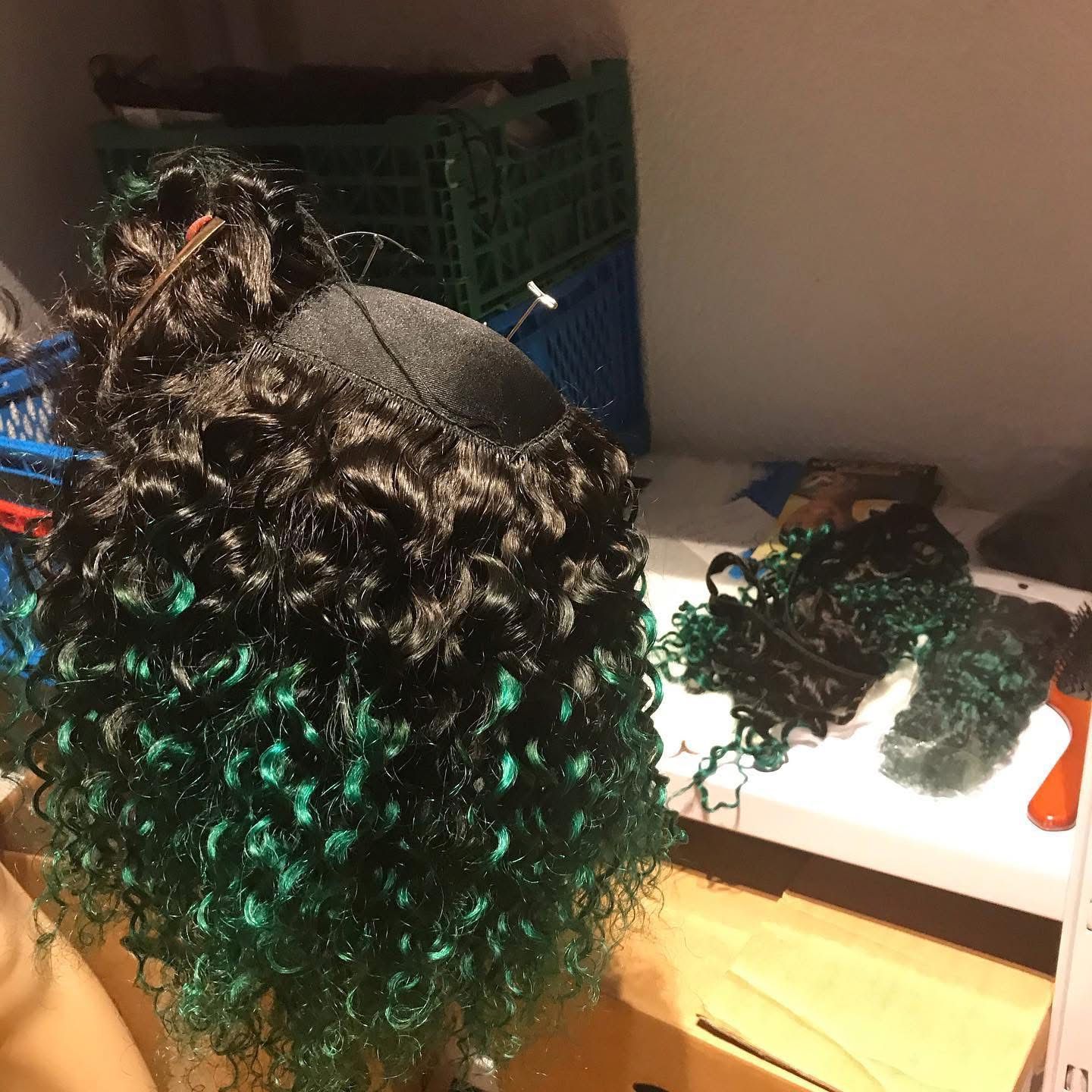 Eine schwarz-grüne Lockenperücke auf einer Kopfform, mit losen Haarsträhnen auf einer weißen Oberfläche daneben.