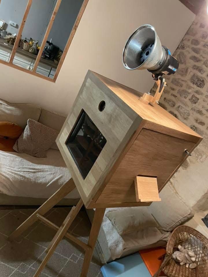 Photobooth en bois incliné avec une fenêtre et un spot, dans une pièce confortable avec un canapé et un miroir mural.