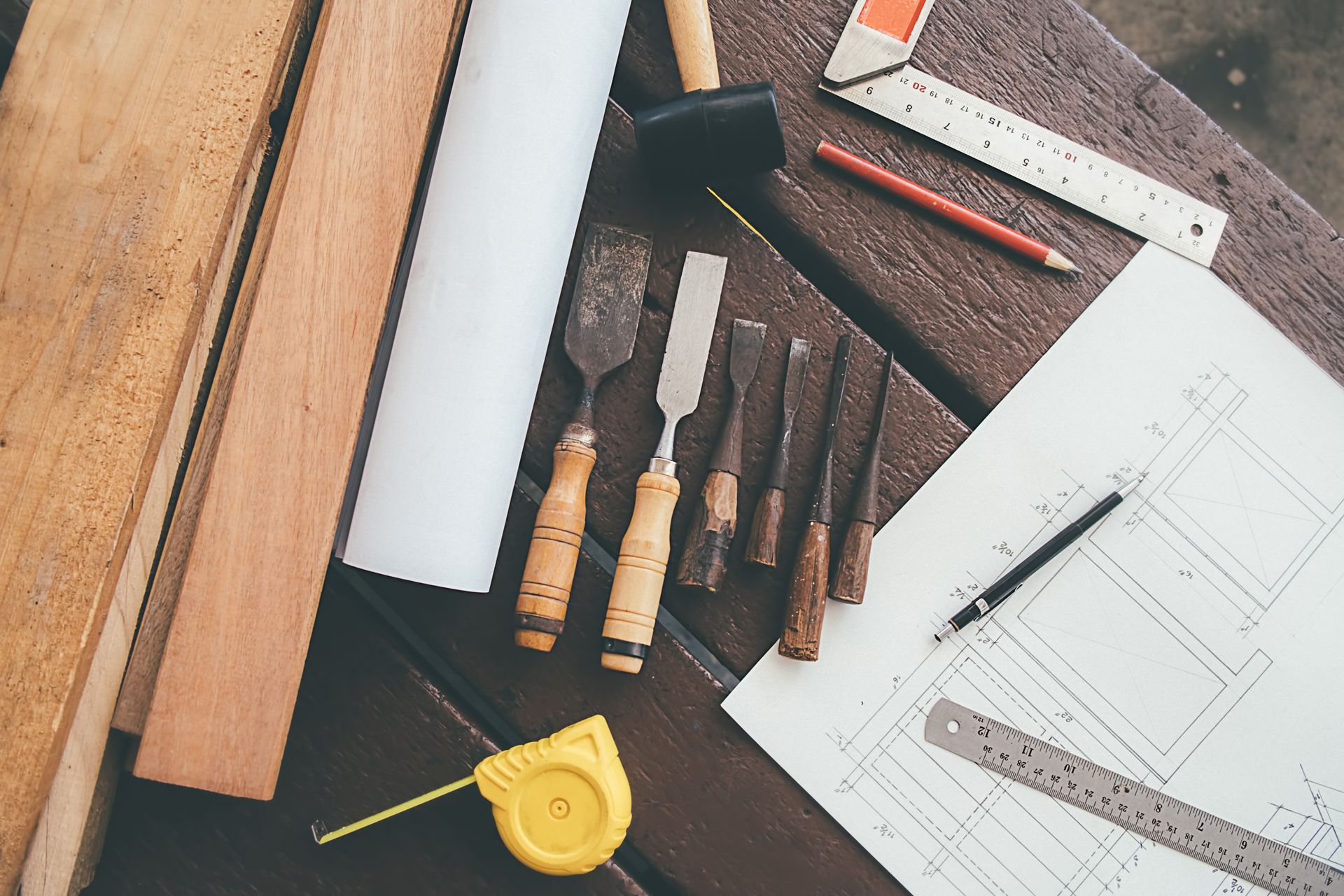 Outils de dessin et plans sur un établi en bois, notamment règles, crayons, mètre ruban et ciseaux à bois.