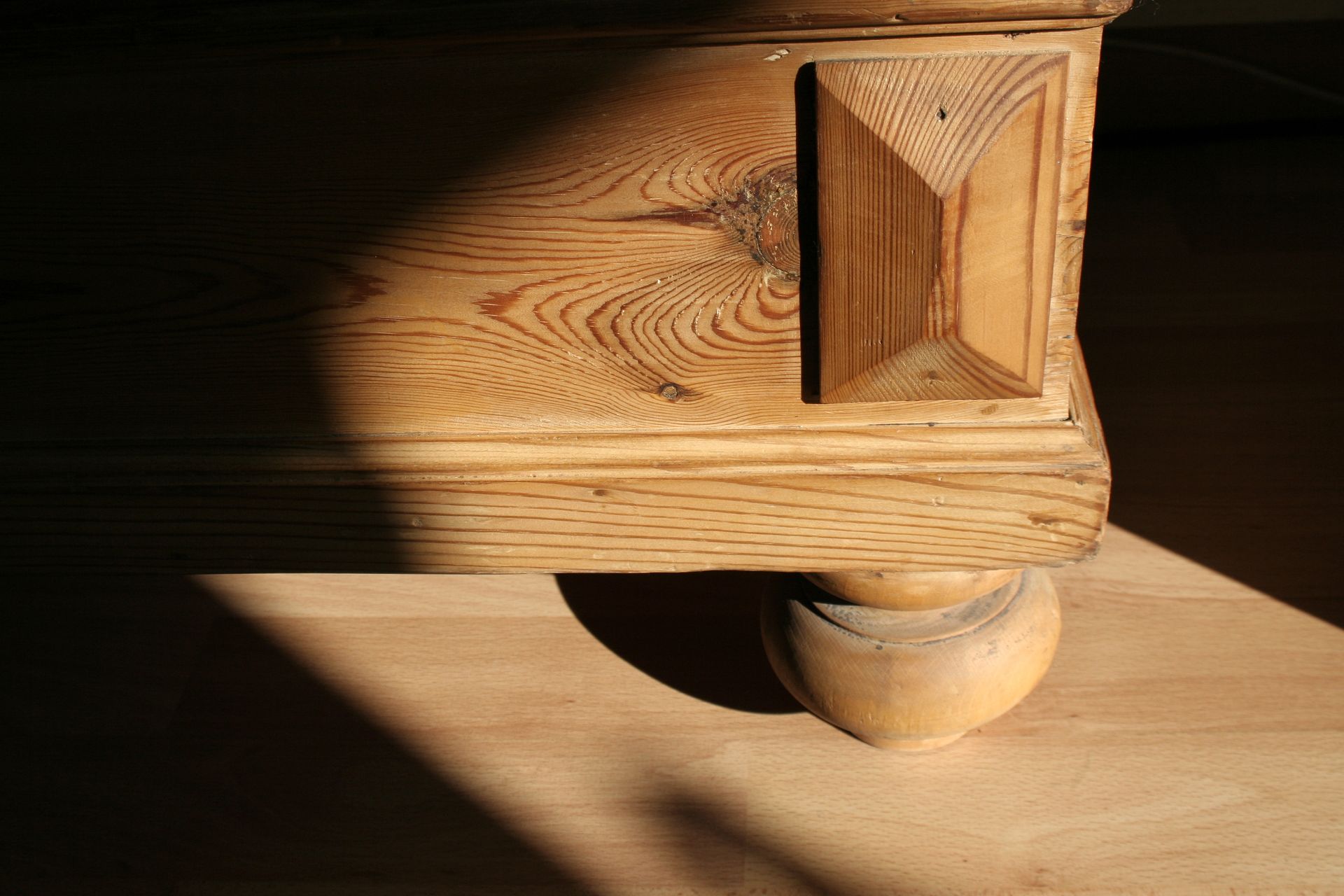 Pied d'un meuble en bois avec une roulette, éclairé par la douce lumière du soleil sur le sol.