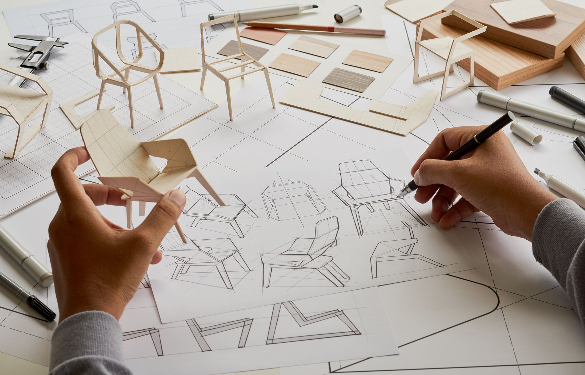 Des mains dessinent des modèles de chaises et assemblent des prototypes de petits meubles en bois sur une table de travail.