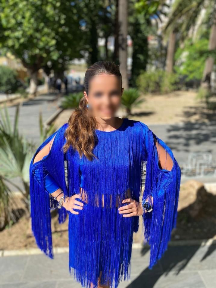 Mujer con un vestido azul con flecos, con los brazos en las caderas, al aire libre en un entorno similar a un parque.