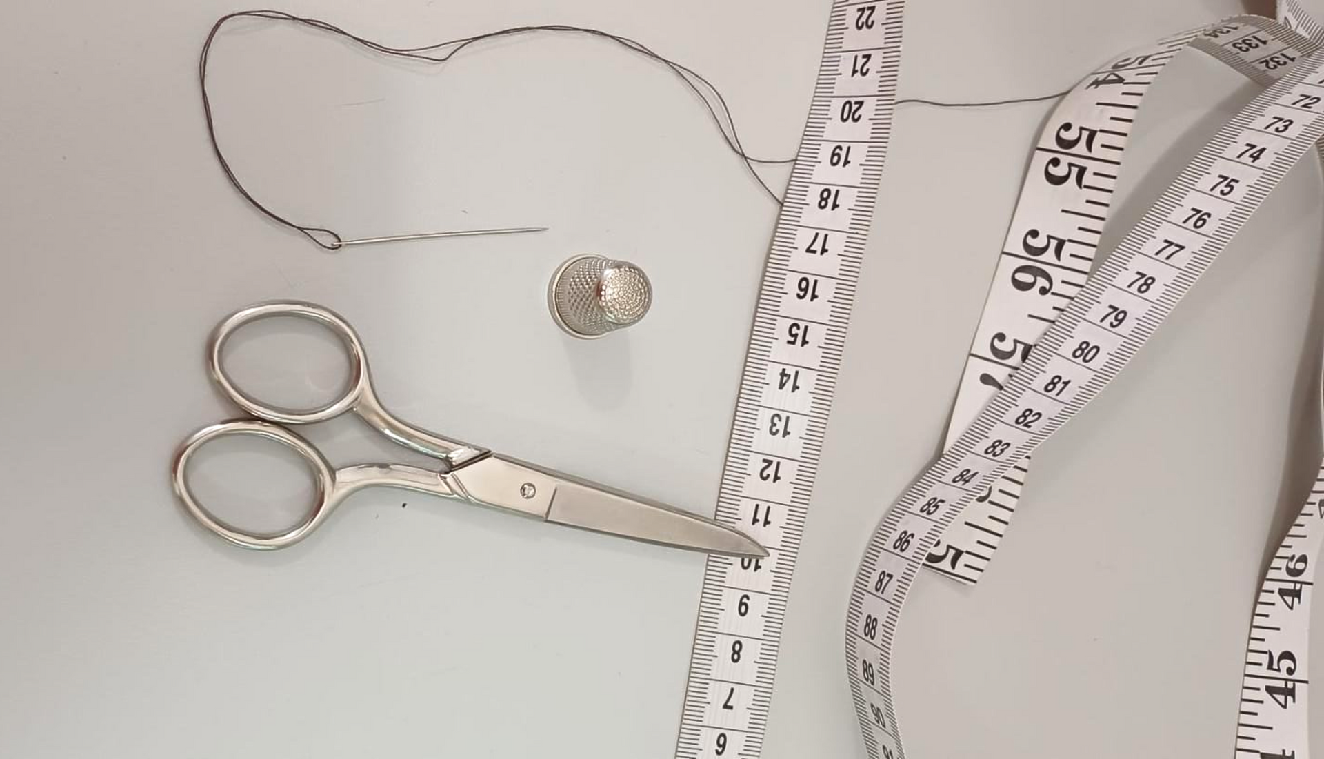 Tijeras, aguja, hilo, botón y cinta métrica sobre una superficie blanca.