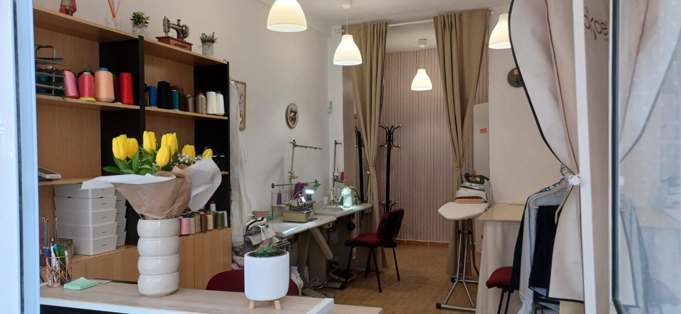 Interior de una sastrería con máquinas de coser, carretes de hilo, tela y flores amarillas.