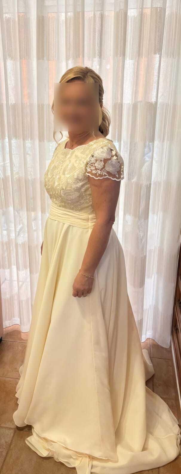 Mujer con un vestido de novia de color crema y mangas cortas, de pie frente a unas cortinas transparentes.