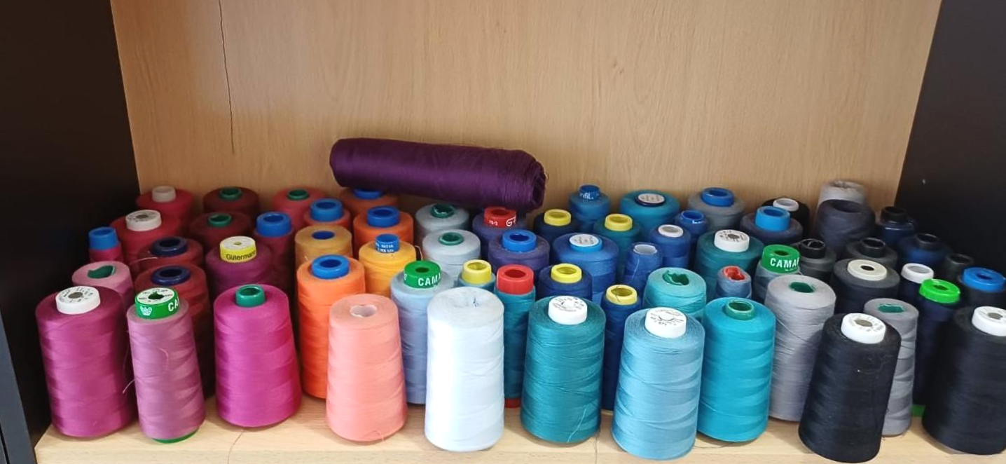 Estante lleno de carretes de hilo de colores: morado, rosa, naranja, blanco, verde azulado, negro y azul.