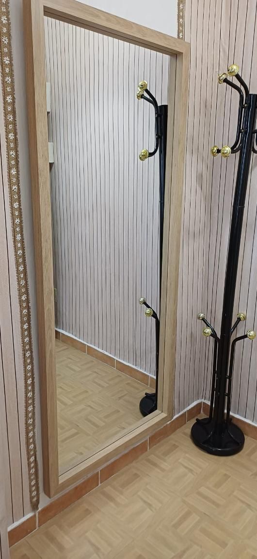 Un espejo de cuerpo entero con marco de madera clara en una habitación. Junto al espejo hay un perchero negro.