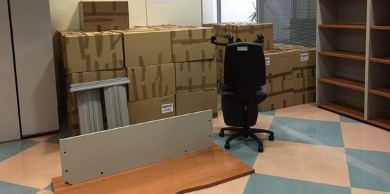 Un espacio de oficina con cajas, piezas de escritorio, una silla y estantes vacíos sobre un piso cuadriculado.