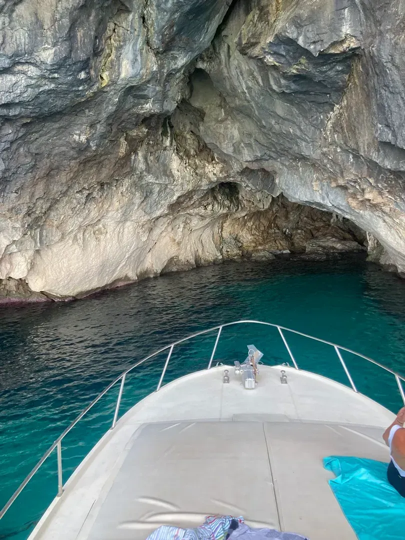 Un barco se acerca a la entrada de una cueva rocosa con agua turquesa y una proa blanca en primer plano.