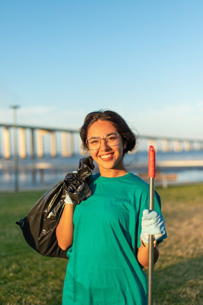 Una persona sonríe al aire libre en una orilla cubierta de hierba, sosteniendo un recogedor de basura y una bolsa negra cerca de un puente al atardecer.
