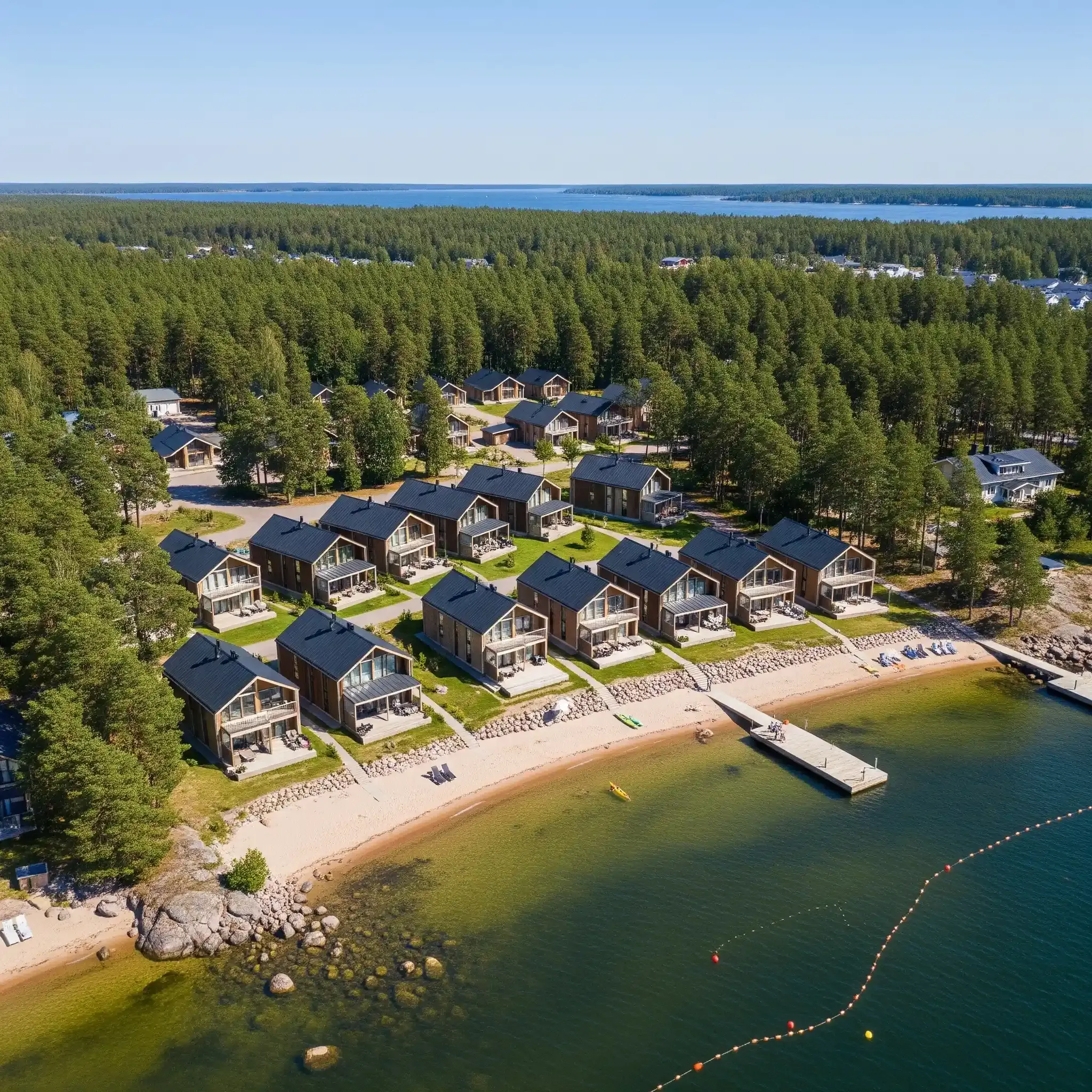 Ilmakuva rantataloista metsän taustalla, laituri ja järvi sinisen taivaan alla.
