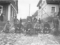 Eine Gruppe von Männern steht und sitzt am Rand einer unbefestigten Straße vor einem Gebäude.
