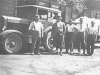 Auf einem Schwarzweißfoto steht eine Gruppe Männer vor einem Lastwagen.