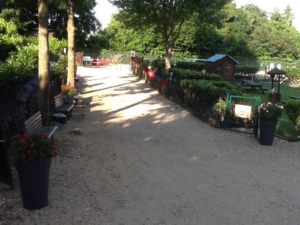 Allée de gravier traversant un jardin bordé de haies, de bancs et de fleurs en pot. Un petit abri de jardin et des tables se trouvent à l'arrière-plan.