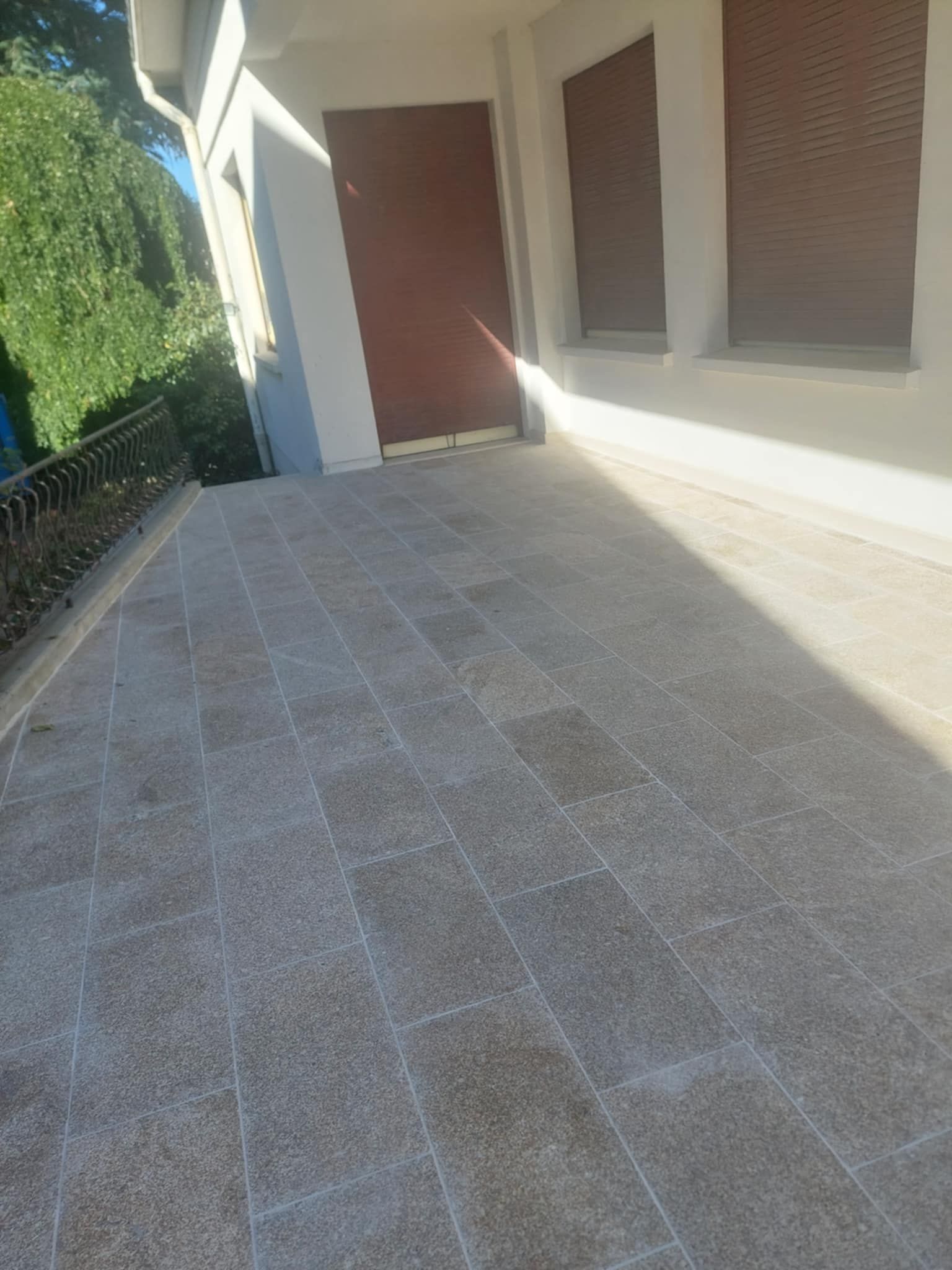 Terrasse en pierre devant un bâtiment blanc aux volets bruns. La lumière du soleil projette des ombres.