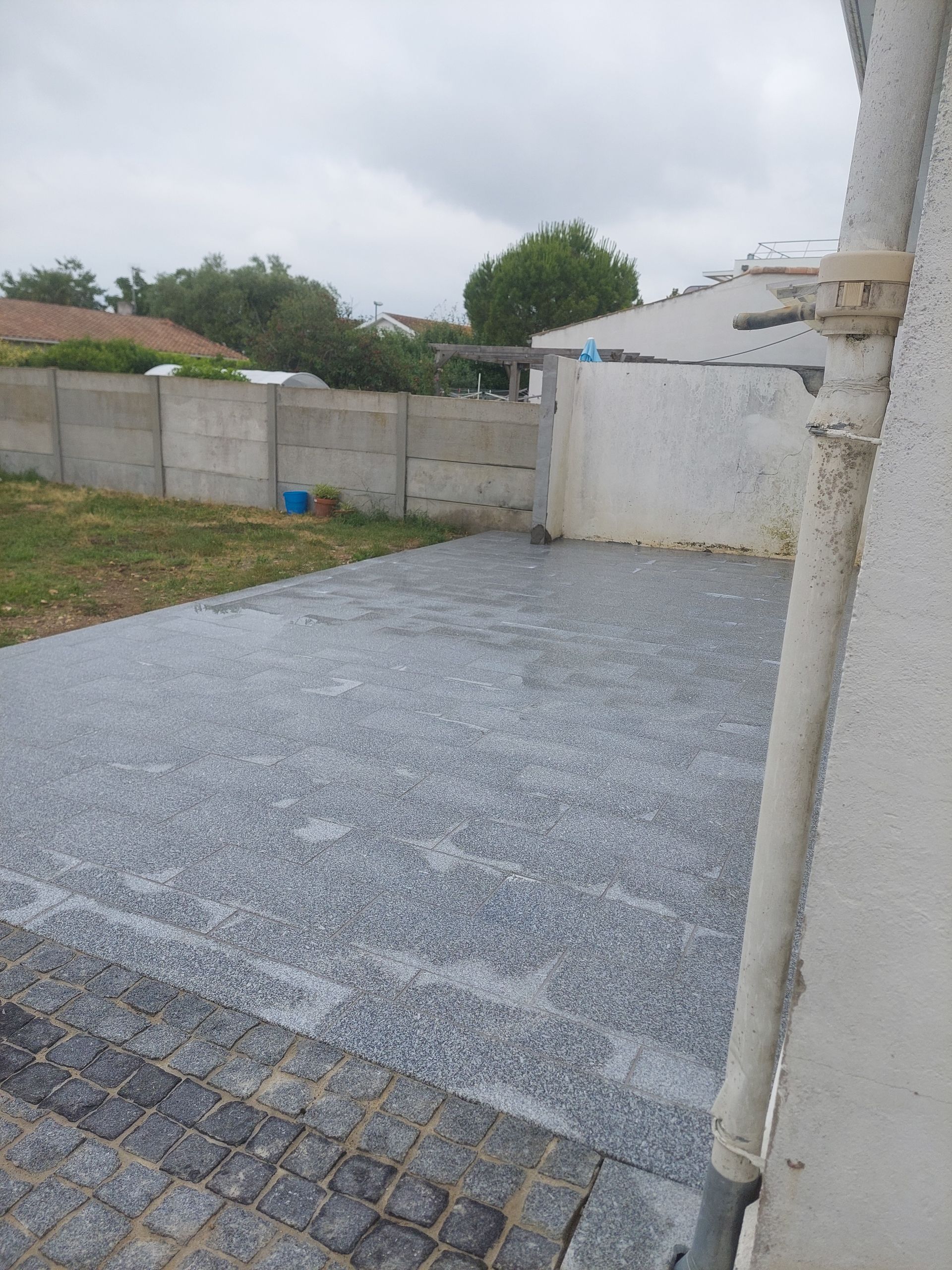 Pluie tombant sur une surface pavée gris foncé dans une cour arrière, avec un mur en béton et un ciel couvert.