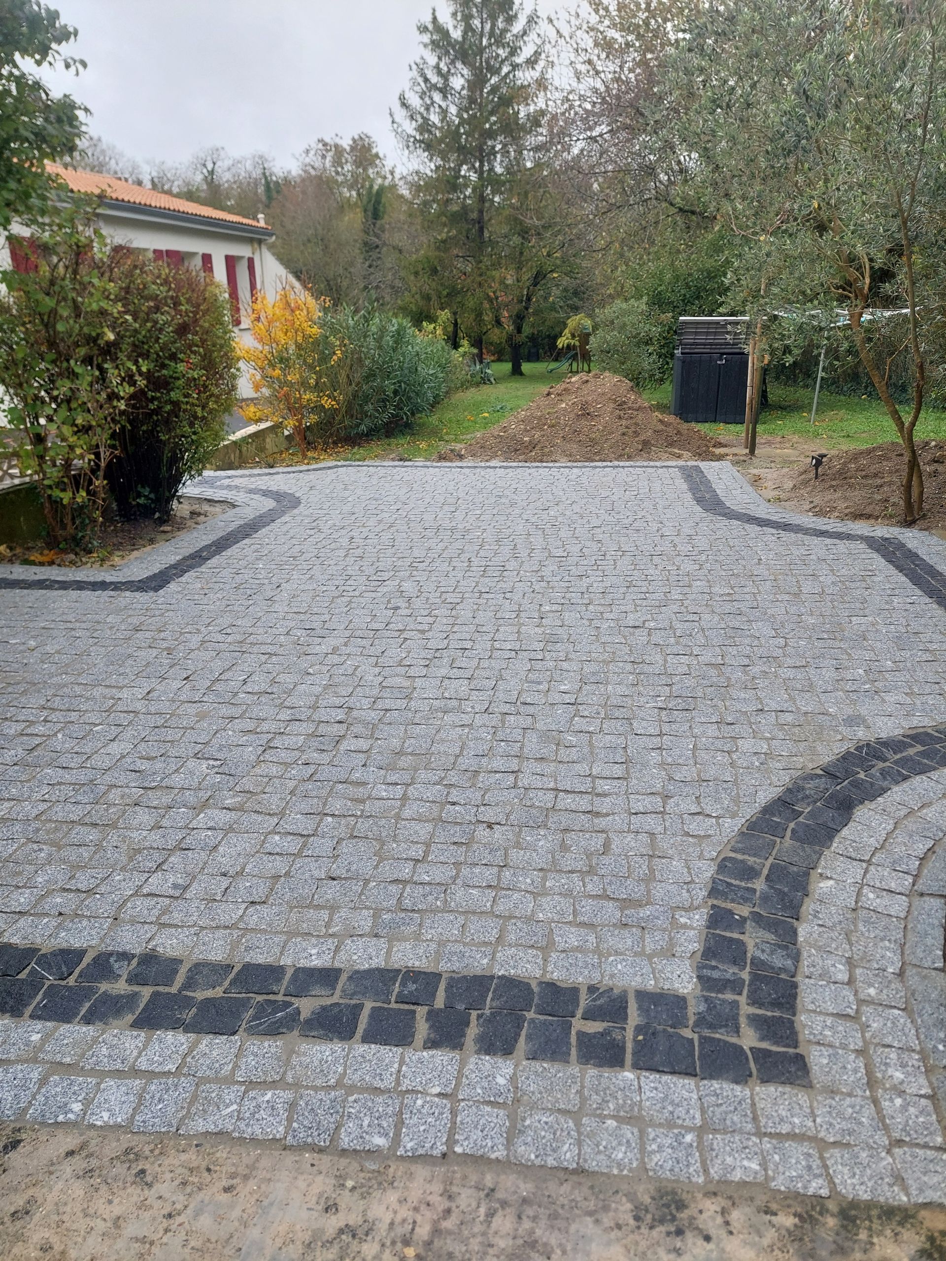 Allée pavée bordée de noir ; une maison et des arbres se trouvent en arrière-plan.