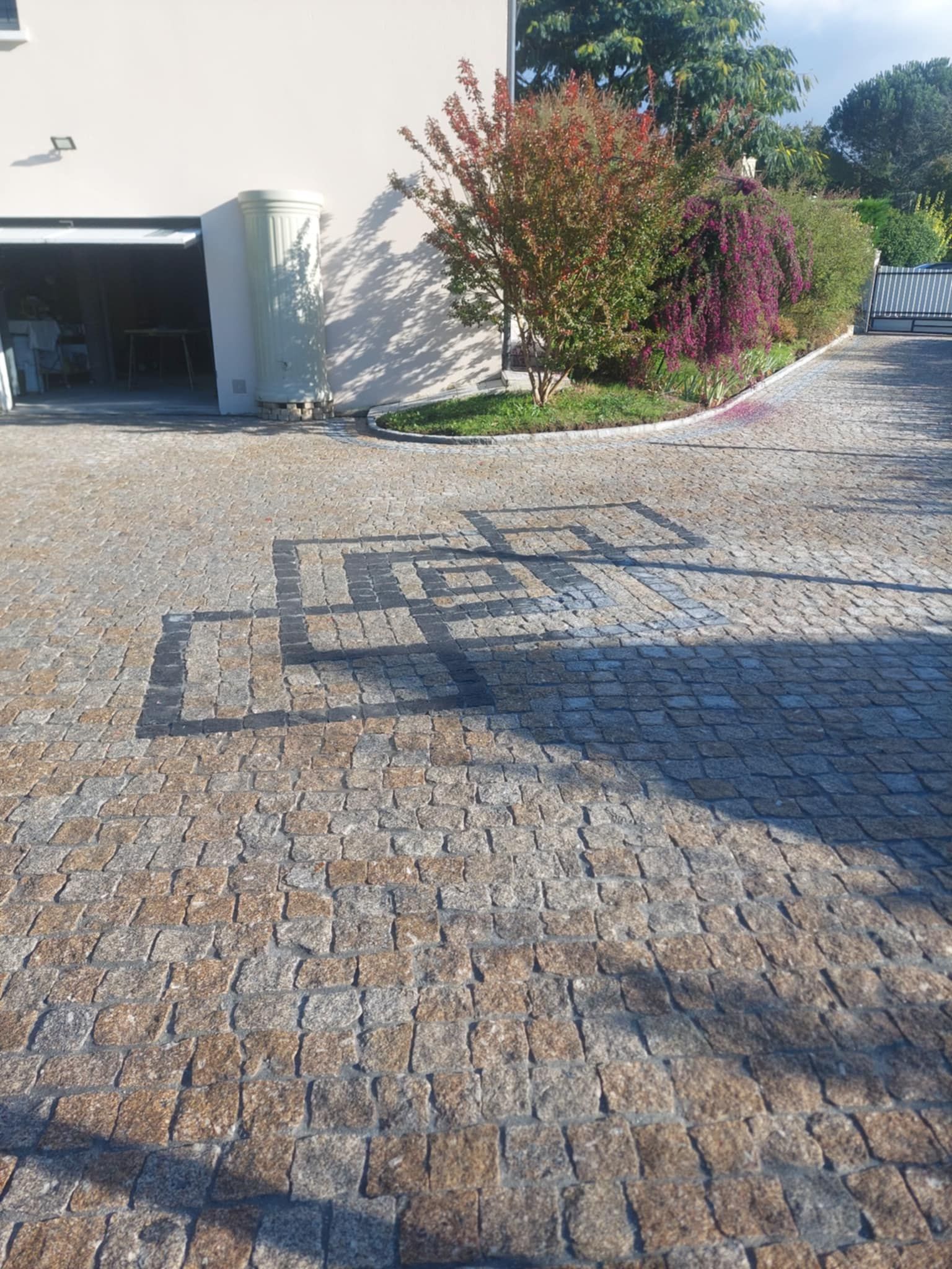 Allée pavée à motifs carrés noirs, jouxtant une maison et un jardin coloré.