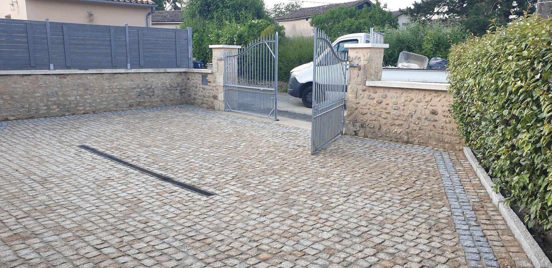 Allée pavée avec portail ouvert et haies vertes. Une camionnette blanche est partiellement visible.