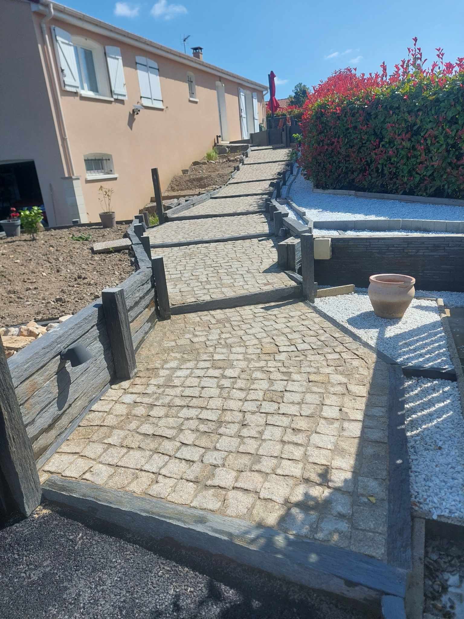 Un chemin pavé bordé de bois grimpe à flanc de colline, longeant une maison et des aménagements paysagers.