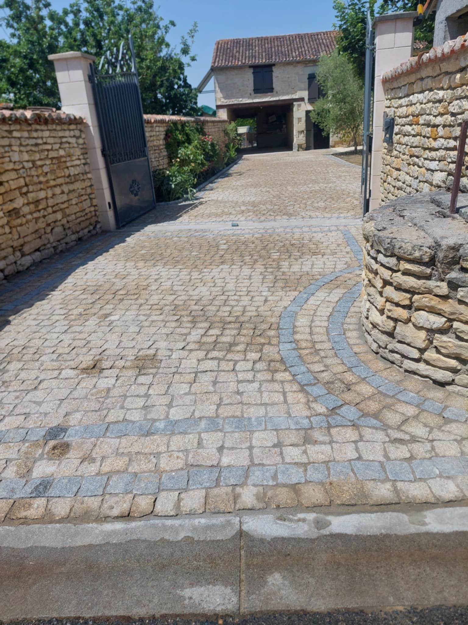 Allée pavée menant à une maison avec un portail ouvert ; pavée de pierres grises et bordée de murs de briques.