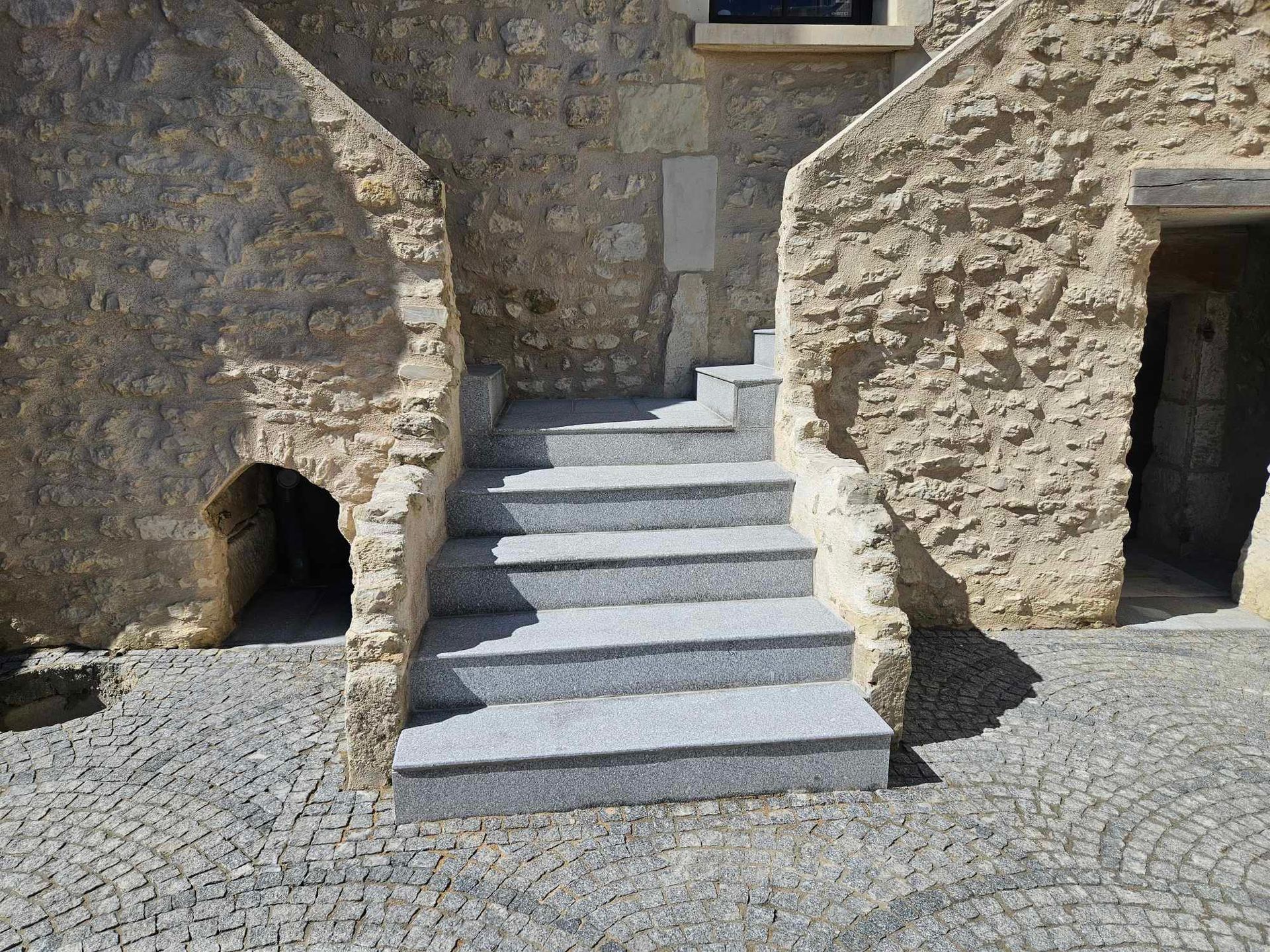Des marches en pierre mènent à l'entrée d'un bâtiment, flanquées de murs en pierre, sol pavé.