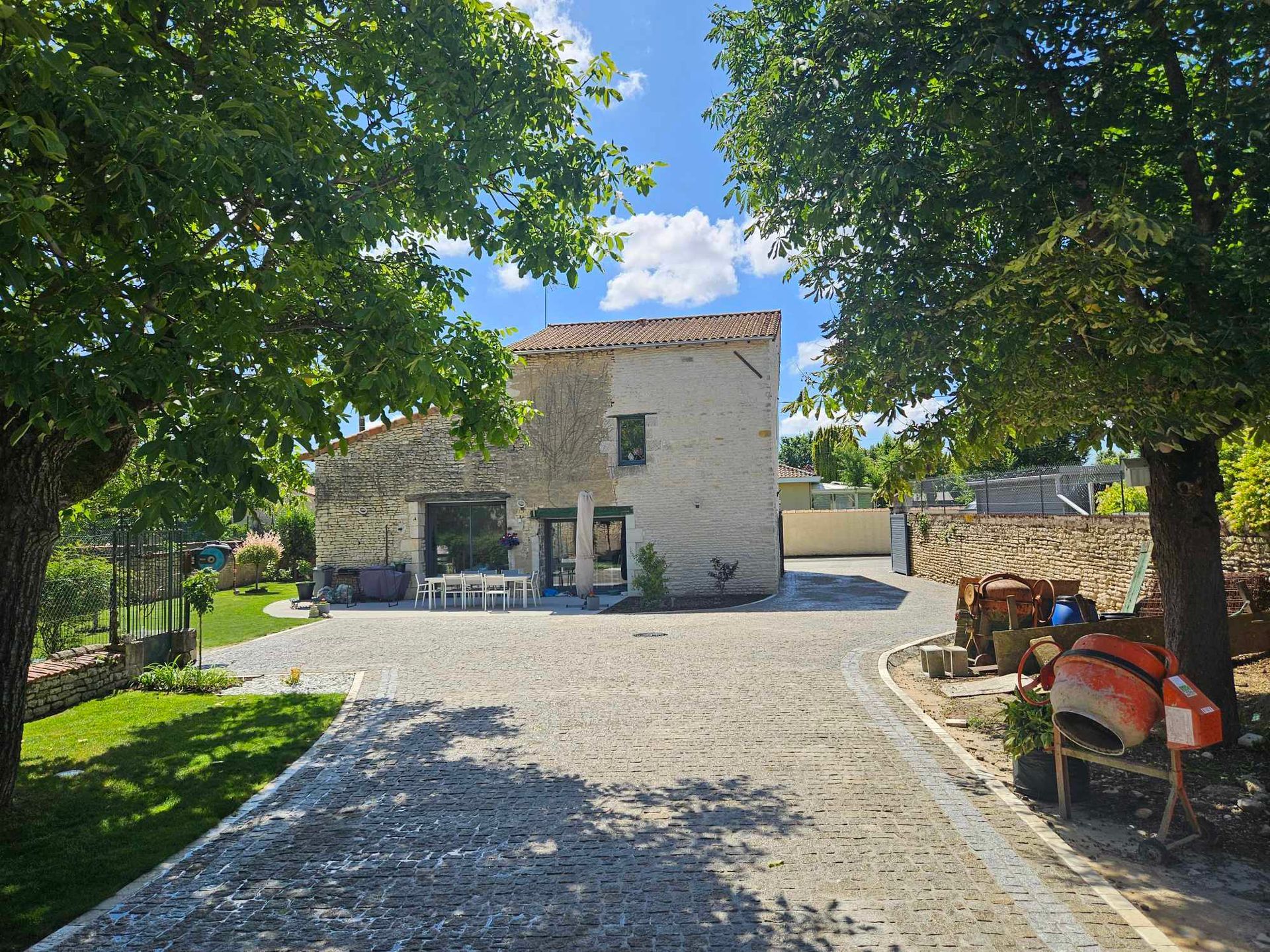Une allée de gravier mène à une maison en pierre, entourée d'arbres sous un ciel bleu.