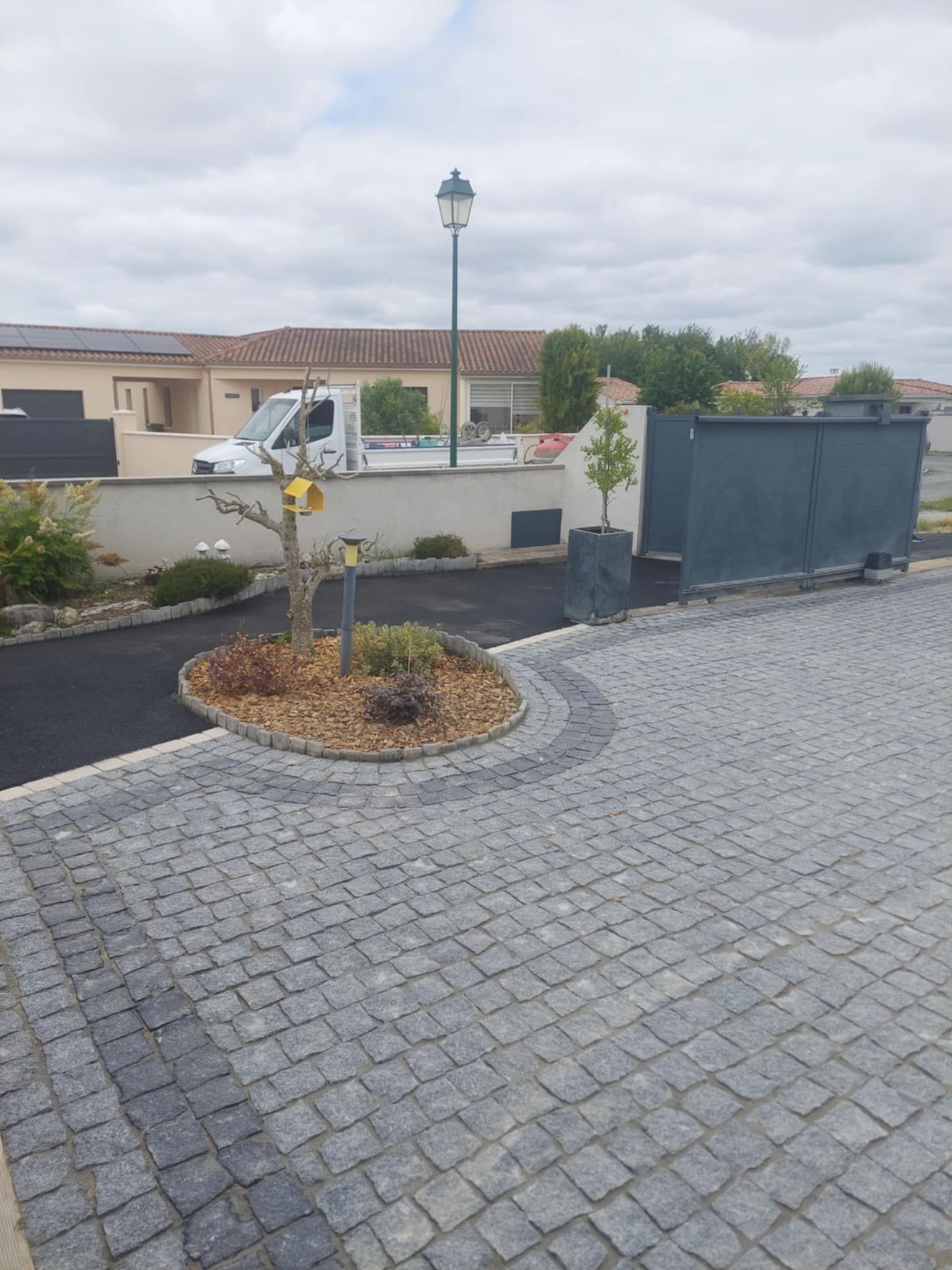 Allée pavée menant à une maison avec un portail noir. Un parterre de fleurs circulaire avec un petit arbre.