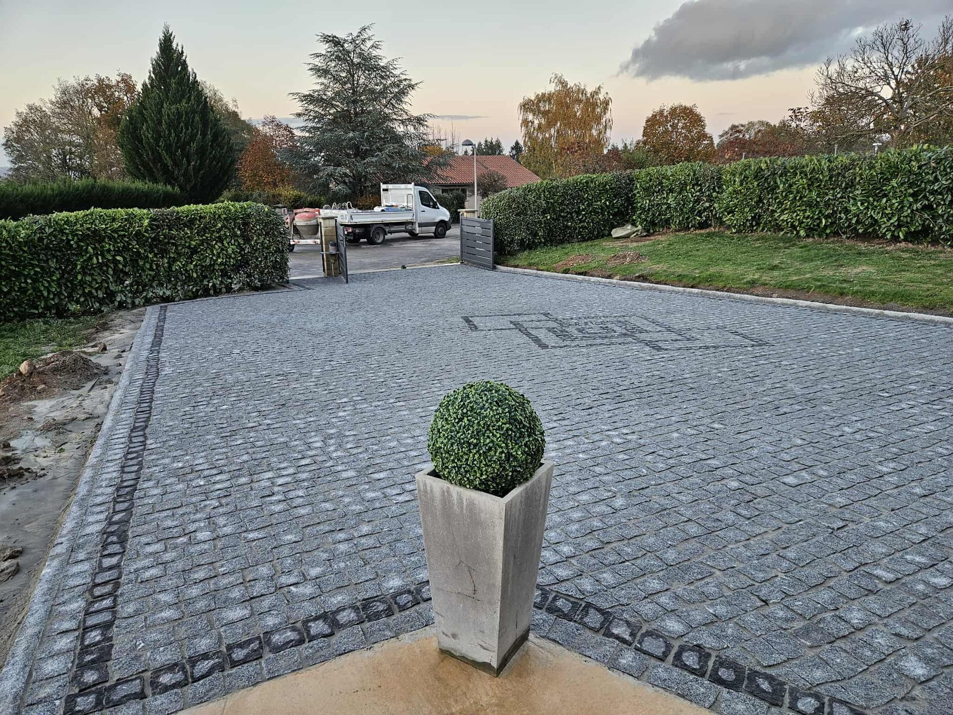 Allée en pierre avec un topiaire dans une jardinière en béton, encadrée de haies et d'arbres.