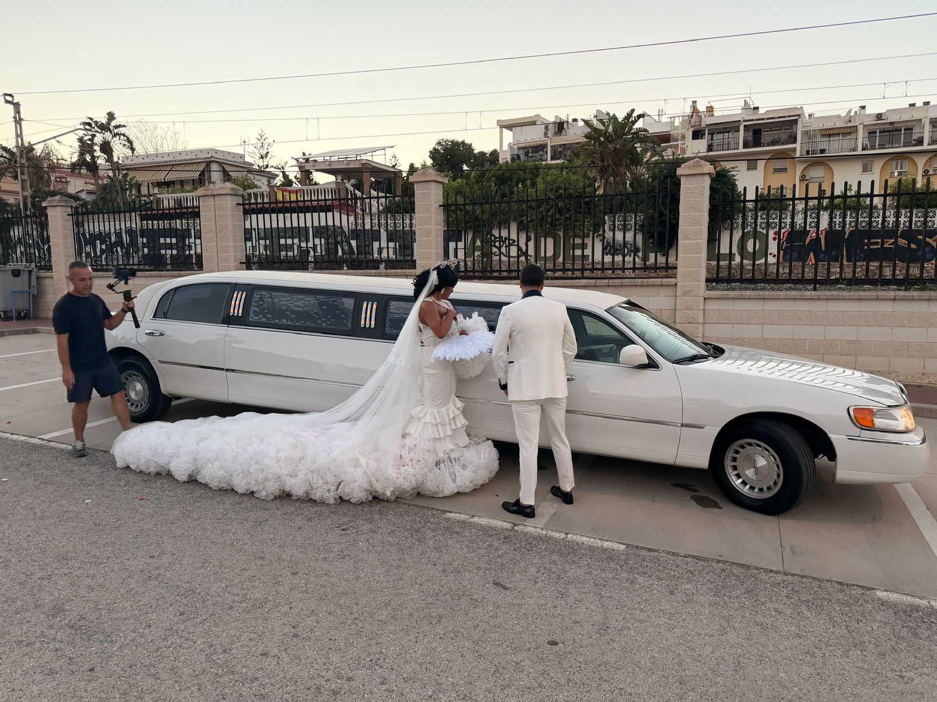 La novia y el novio junto a una limusina blanca alargada en una calle, con la larga cola del vestido de novia