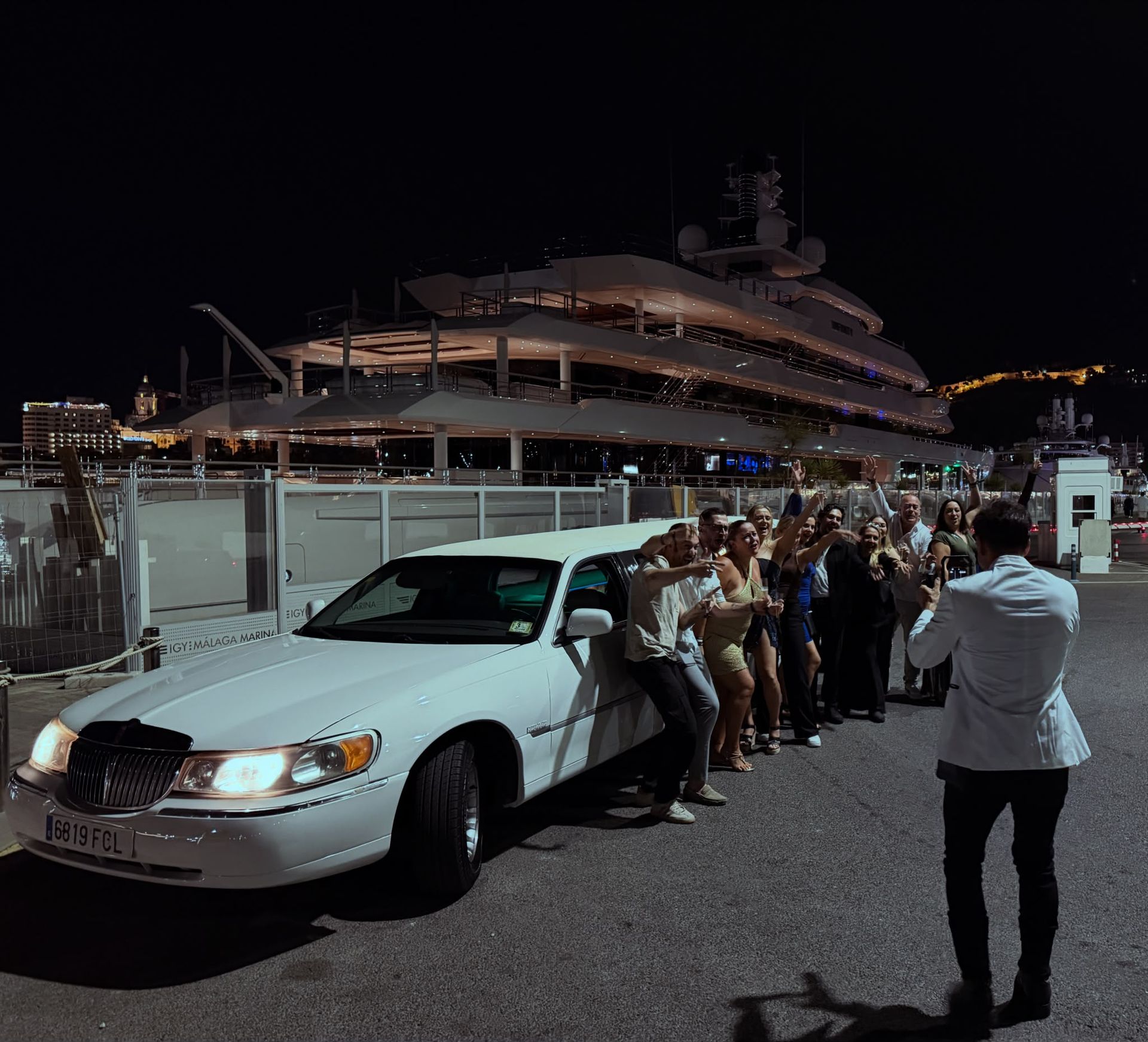 Limusina blanca junto a un yate atracado de noche, con un grupo posando y un fotógrafo cerca.