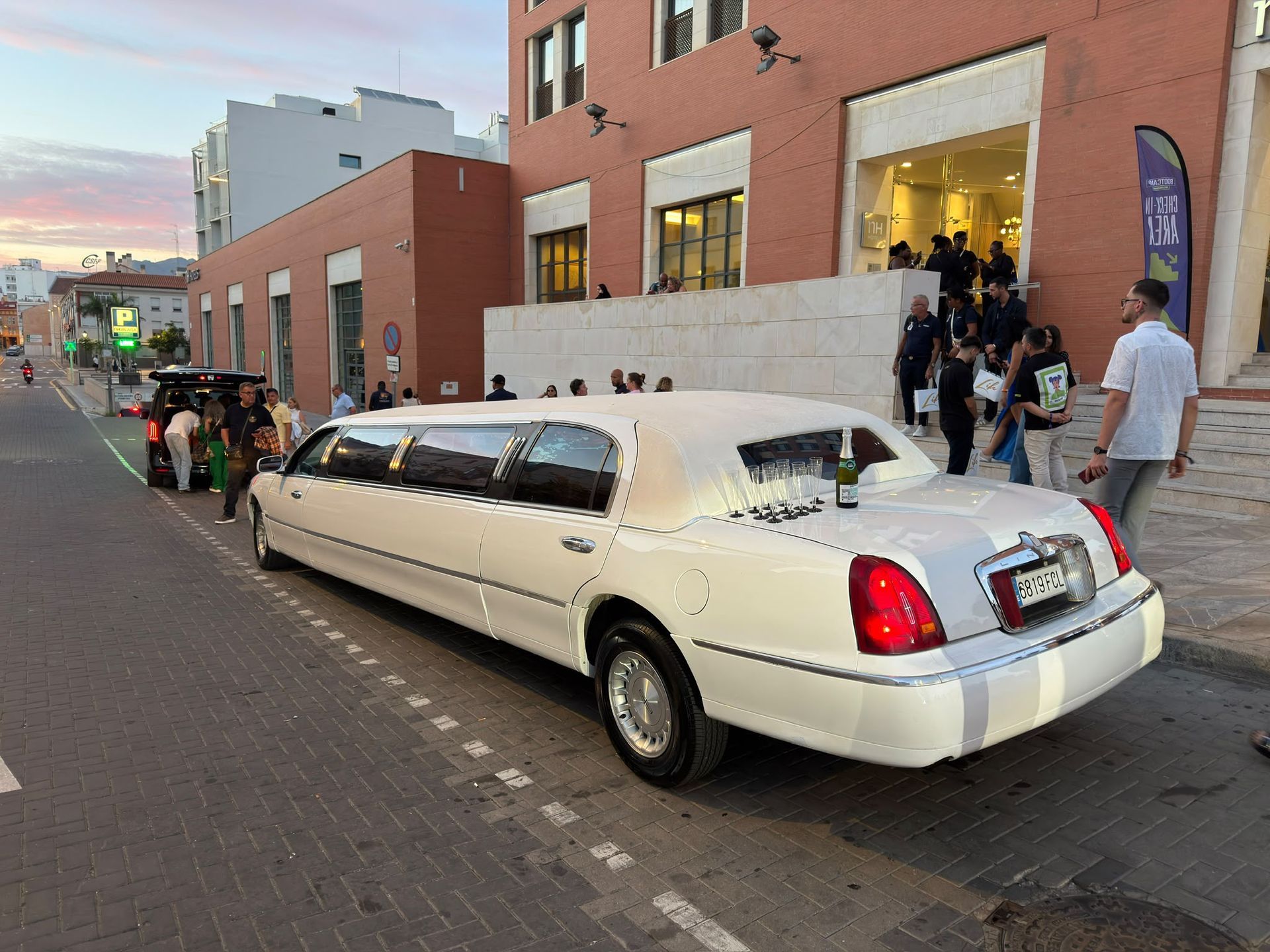 Limusina blanca alargada estacionada en una calle de la ciudad, junto a un edificio de ladrillo con gente reunida en las escaleras.