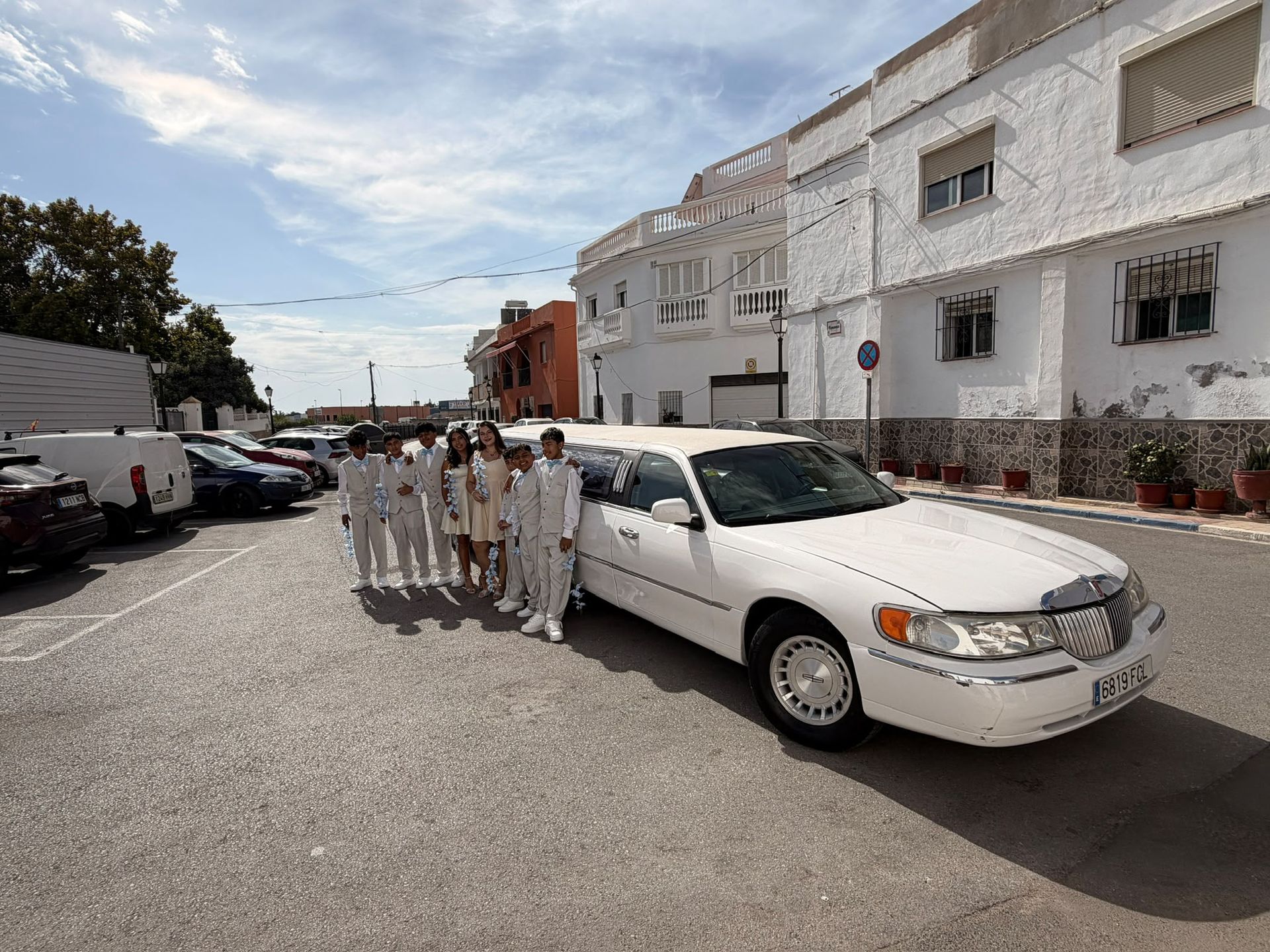Unas personas vestidas de blanco están junto a un coche blanco en una calle soleada con edificios blancos.