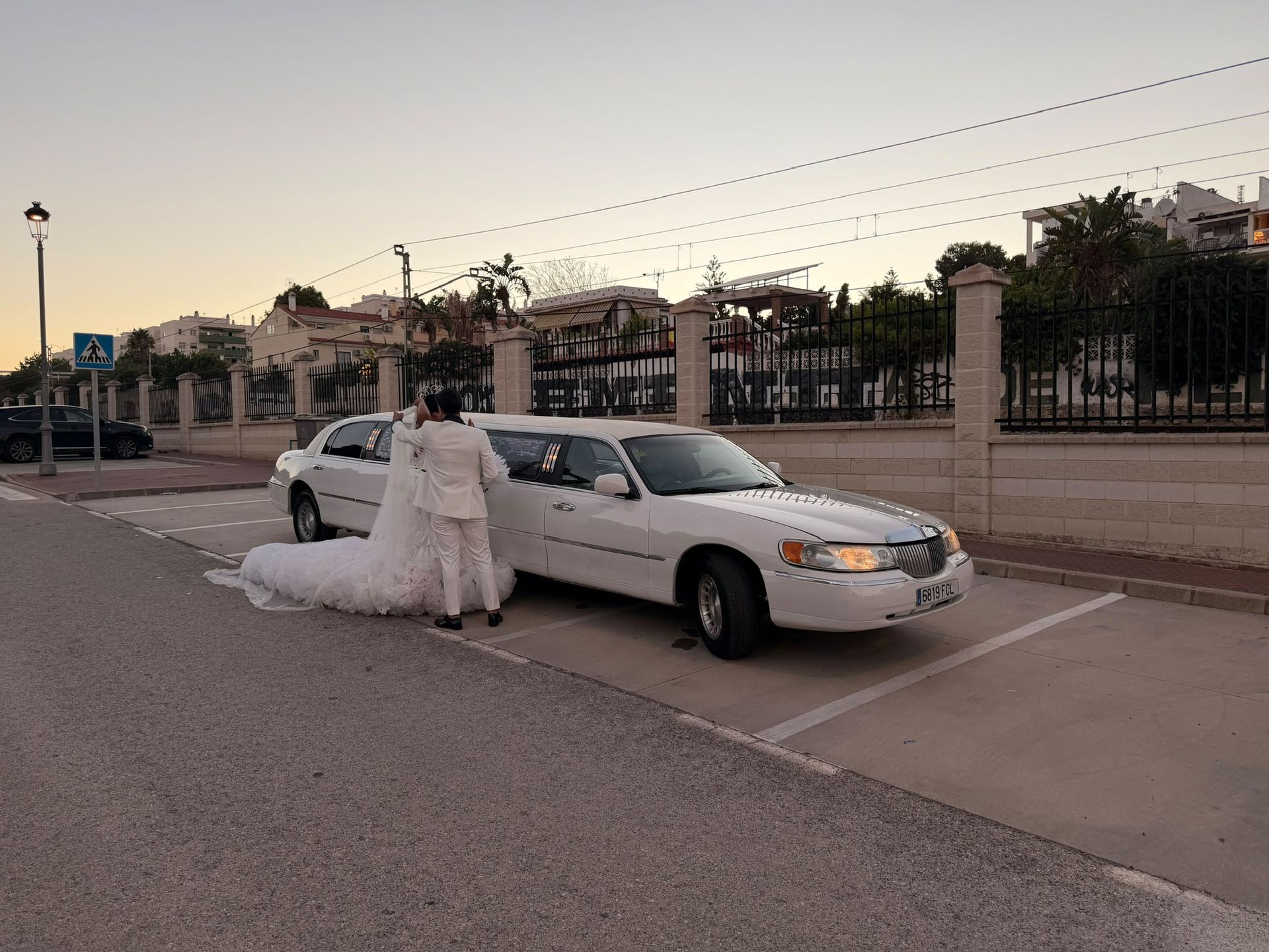 Novia con vestido blanco apoyada en una limusina blanca en una calle tranquila al anochecer