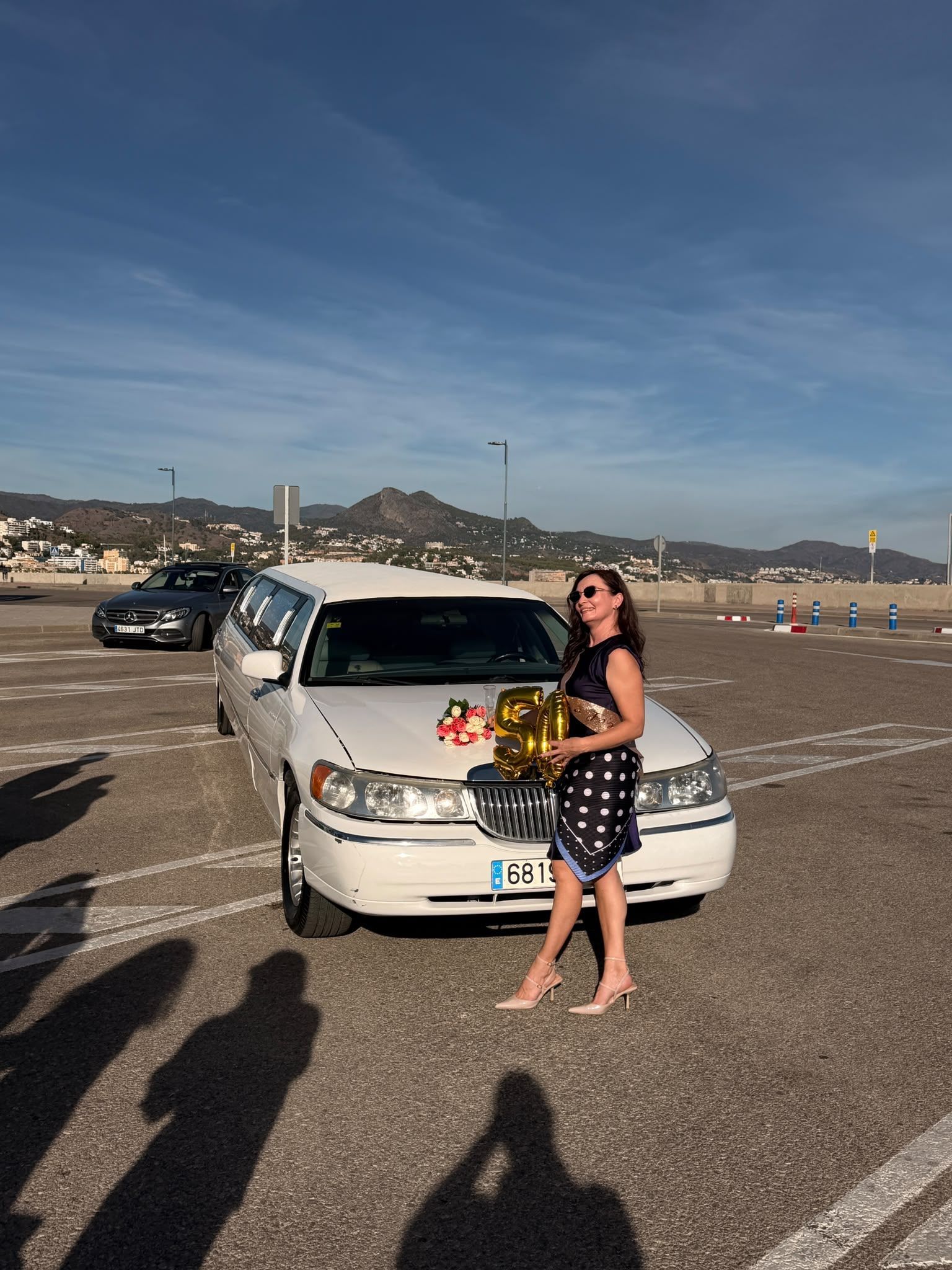Mujer de pie junto a un coche blanco con flores en una carretera costera y montañas al fondo