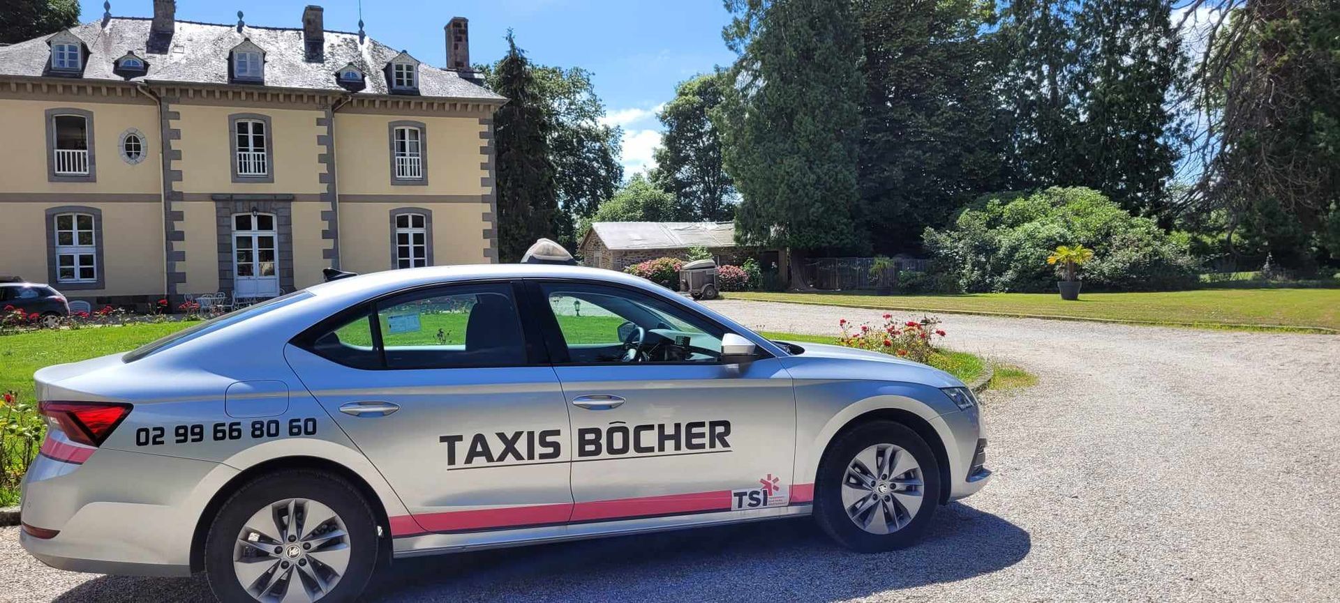 Taxi blanc, floqué de l'inscription Taxis Bôcher, garé devant une maison beige, dans une allée ensoleillée avec des arbres.