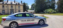 Un taxi argenté garé à côté d'un bâtiment en pierre dans une cour ensoleillée, avec des arbres et du gravier