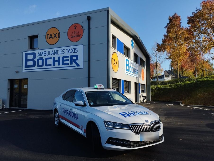 Un taxi blanc est garé devant un bâtiment portant l'enseigne Ambulances Taxis Bôcher