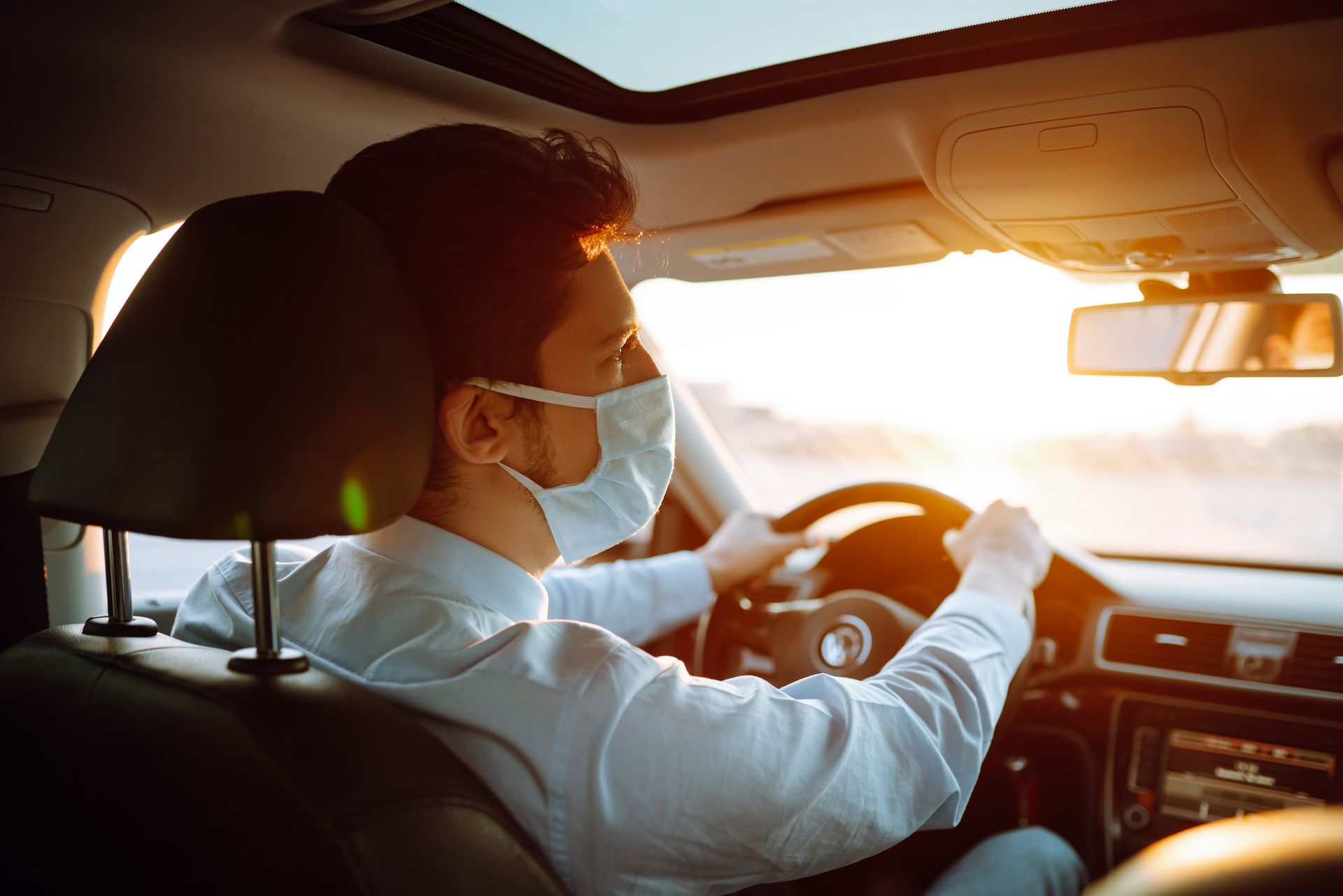 Un conducteur masqué conduit une voiture sous un coucher de soleil.